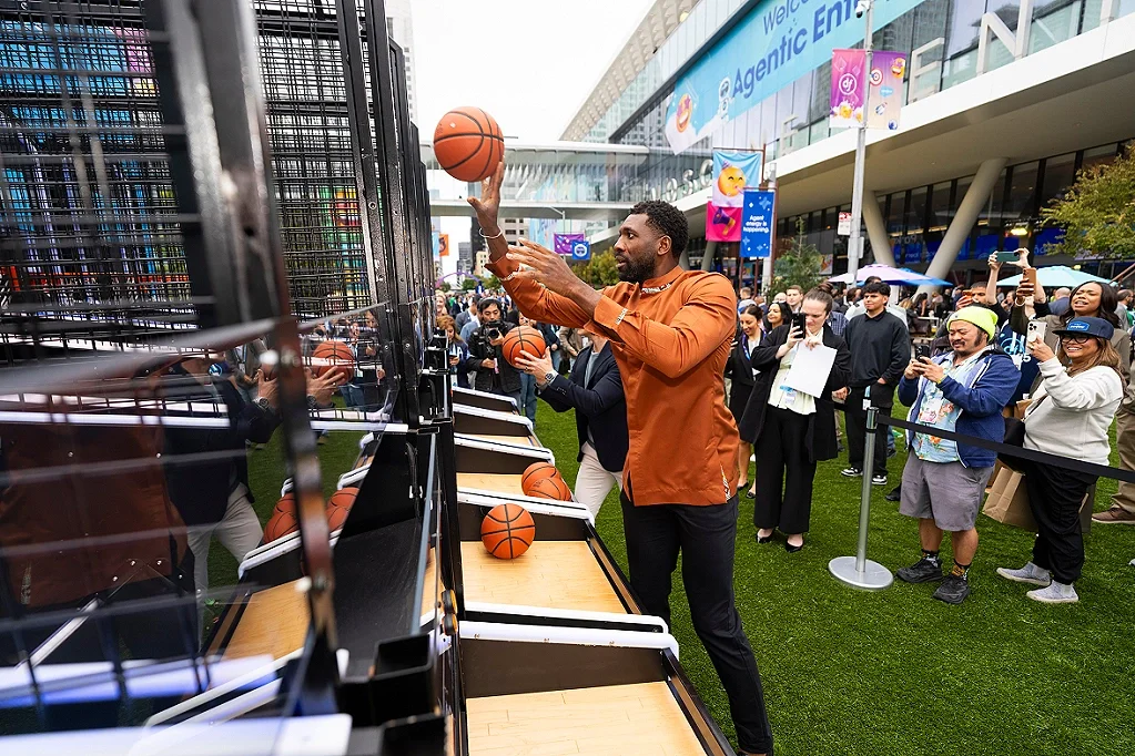 A man in an orange shirt playing basketball on a vertical hoop game at an outdoor event, with a crowd of people watching and taking photos in front of a modern building with a large blue banner.