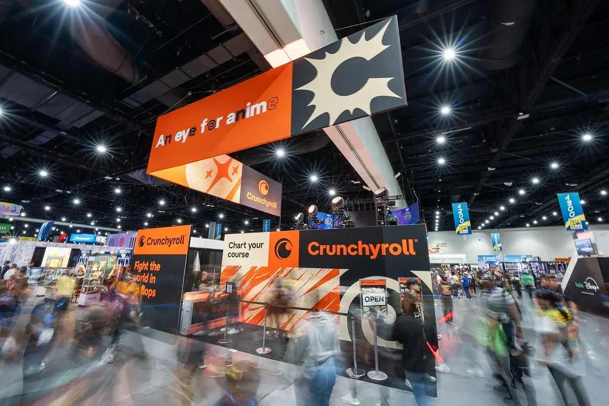 Trade show booth for Crunchyroll featuring large overhead banners and displays, with many visitors walking around the event.