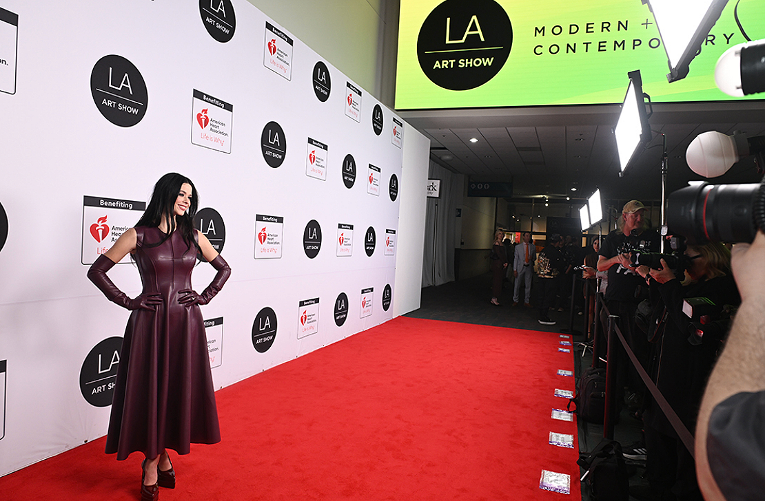A woman in a long, dark purple dress poses on a red carpet at the LA Art Show, with a backdrop displaying the event's logo and sponsors. Photographers and camera operators are capturing her photo.