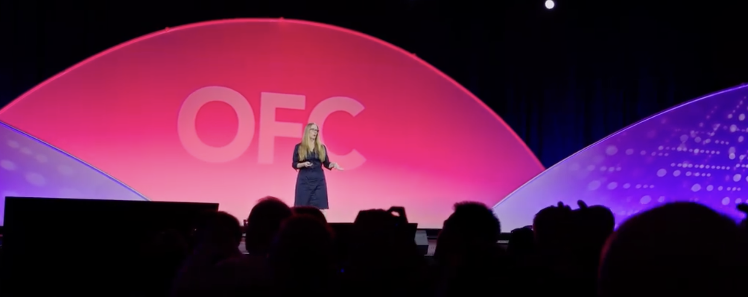 A woman in glasses and a long dark dress speaking on stage at a conference, with a large pink and purple abstract backdrop showing the letters 'OFC'.