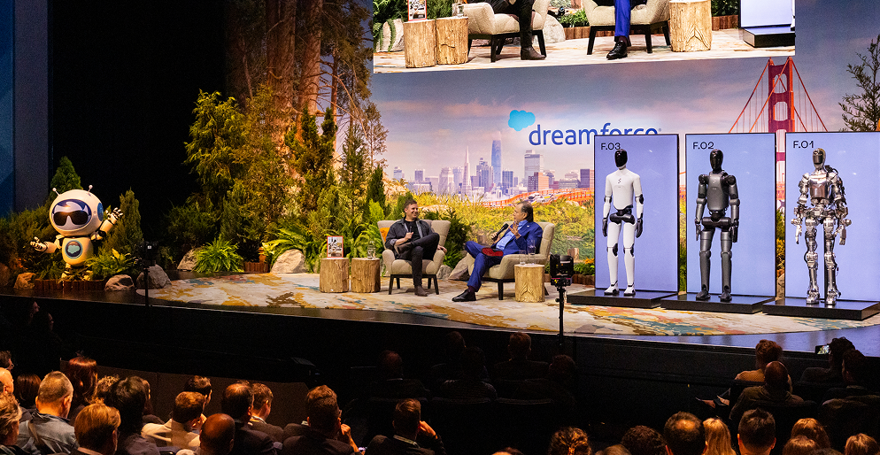 Technological conference or talk with a panel of two men on stage, three robot models labeled F.01, F.02, and F.03, and a backdrop displaying San Francisco and the Golden Gate Bridge, with the logo 'dreamforce.