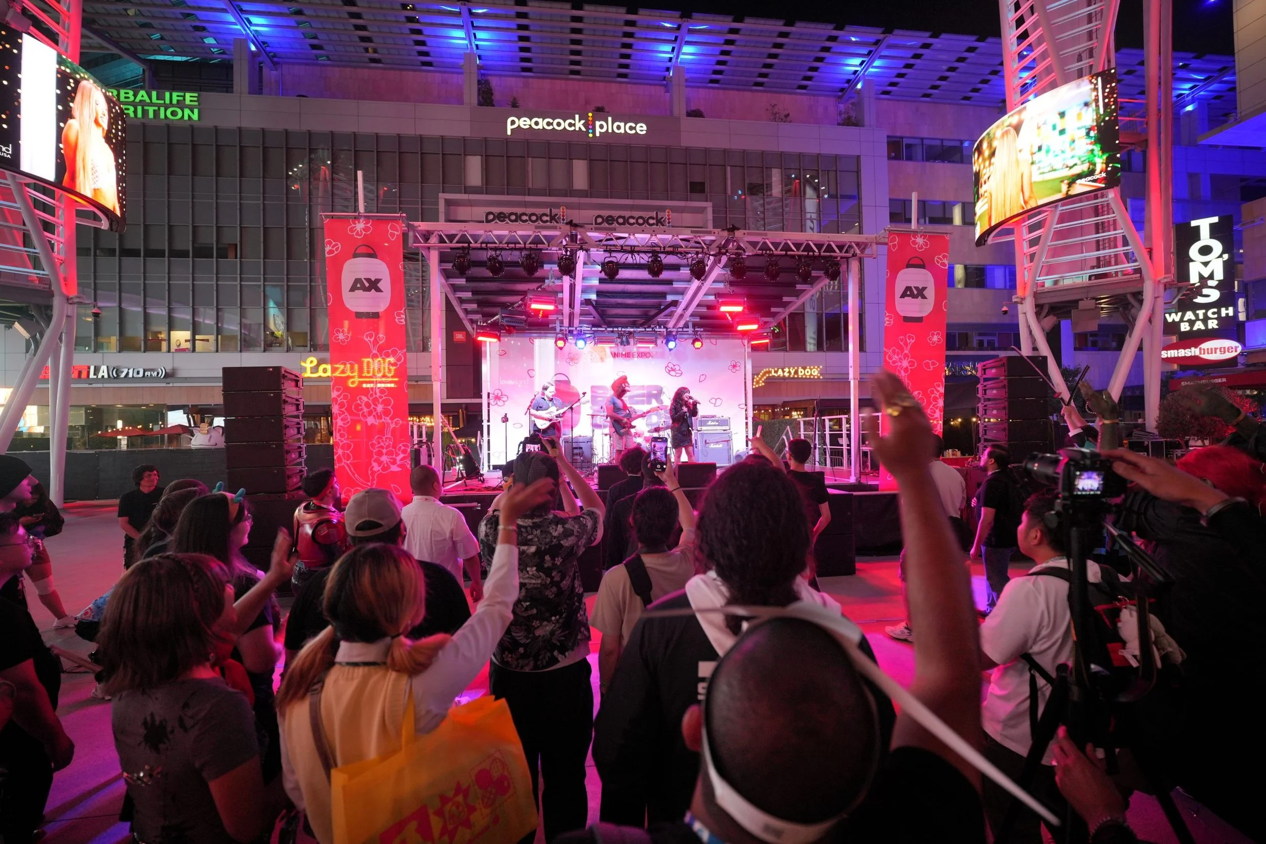 Outdoor concert stage at night in a shopping mall with performers playing guitar and singing, audience taking photos, large digital screens and colorful lights, signage for Peacock Place and other stores.