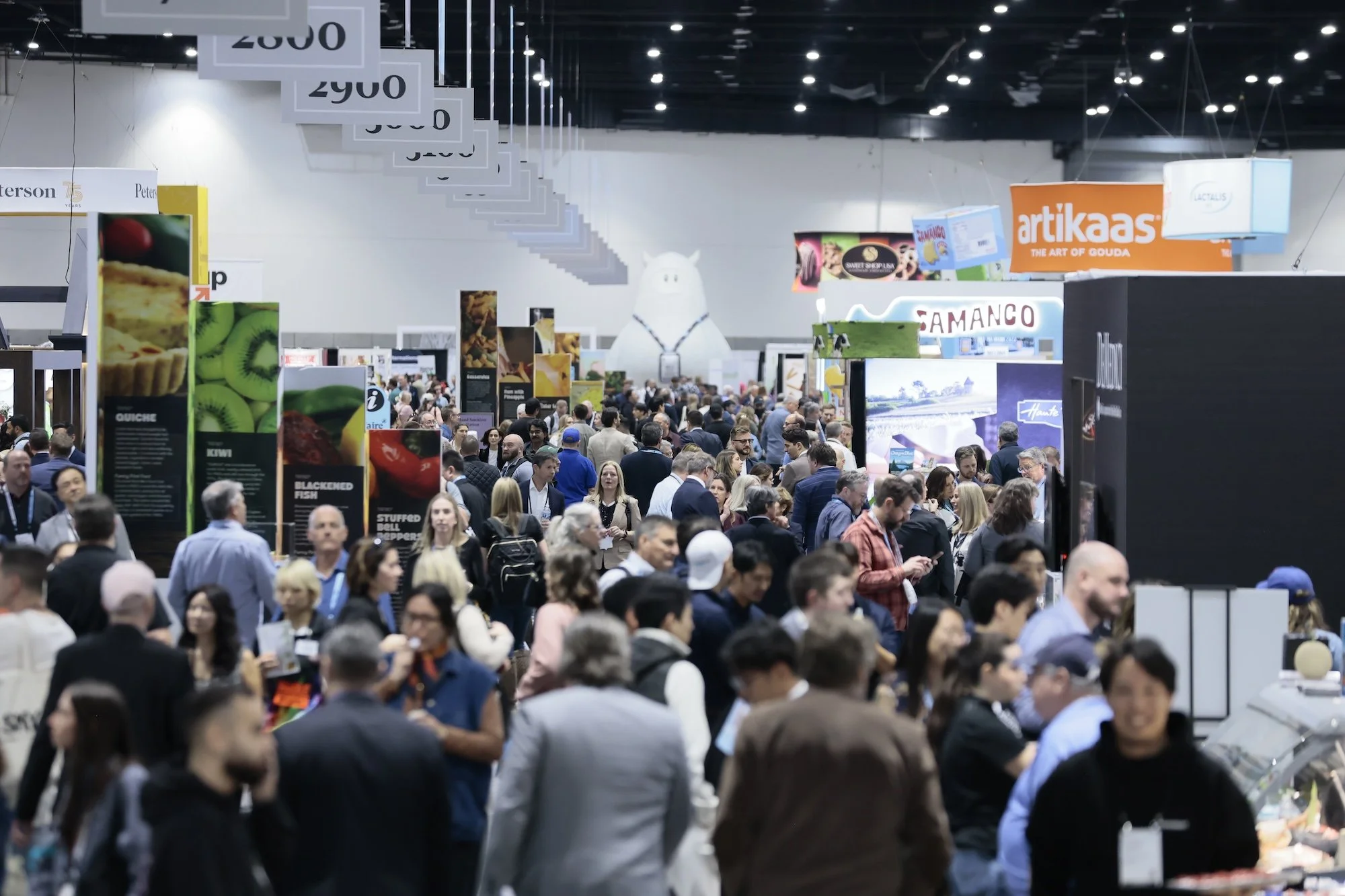 A crowded convention or trade show with numerous booths, signs, and a large inflatable figure resembling a white animal, possibly a cow or a pig, in the background. People are walking and browsing the exhibits.