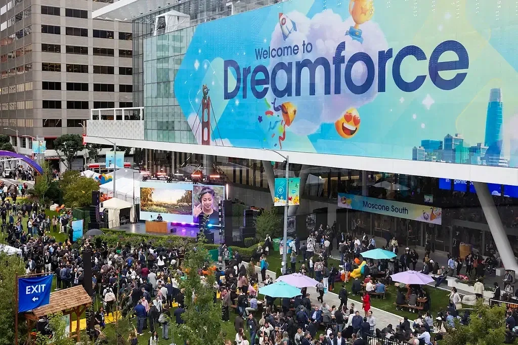 Crowd of people outside at Dreamforce convention with a large digital billboard that says 'Welcome to Dreamforce' and a stage with a speaker.