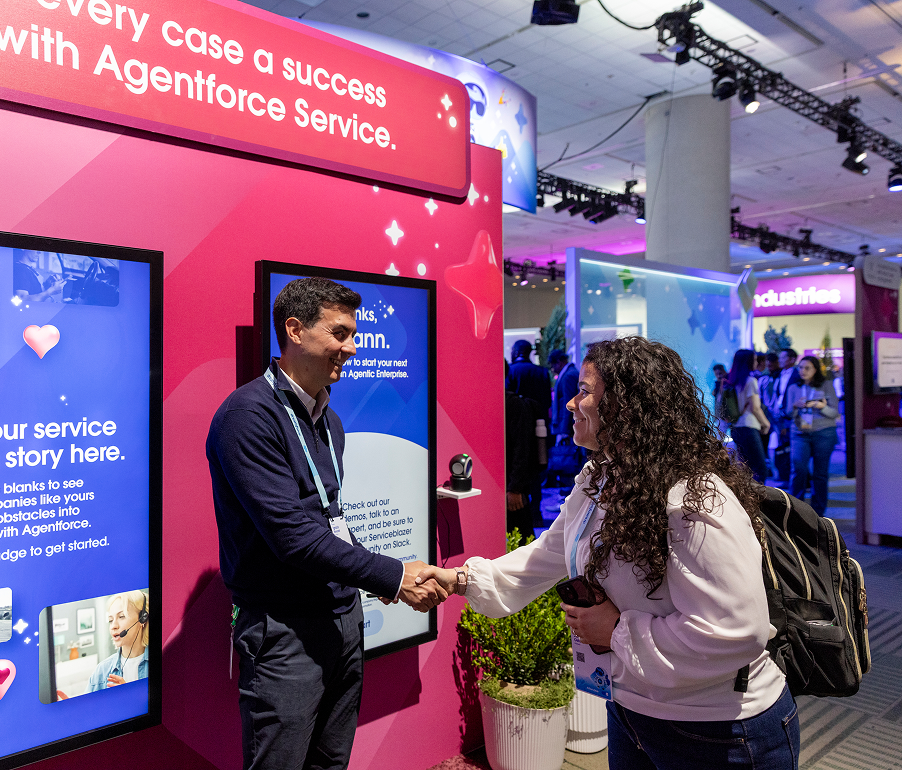 A man and a woman are shaking hands at a professional event or conference, standing in front of a bright pink display with screens showcasing information about a service called Agentforce. The woman has long curly hair, is wearing a white jacket and a backpack, and is holding a camera, while the man has short dark hair and is dressed in a navy blue sweater.