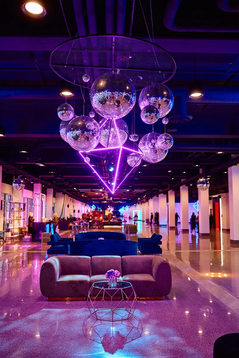 A vibrant, neon-lit lounge with disco balls hanging from the ceiling, a modern sofa and table with flowers, and colorful lighting creating a lively atmosphere.