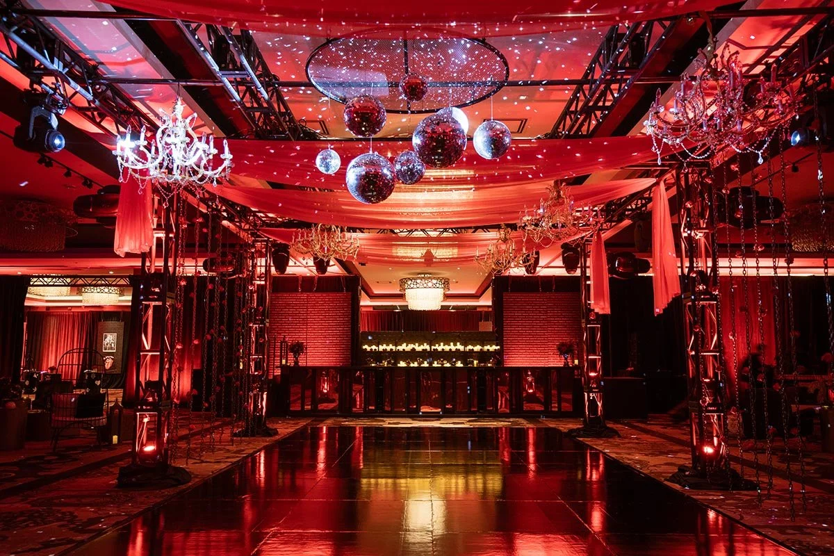 A decorated event space with red lighting, chandeliers, and hanging disco balls, featuring a dance floor and elegant drapery.