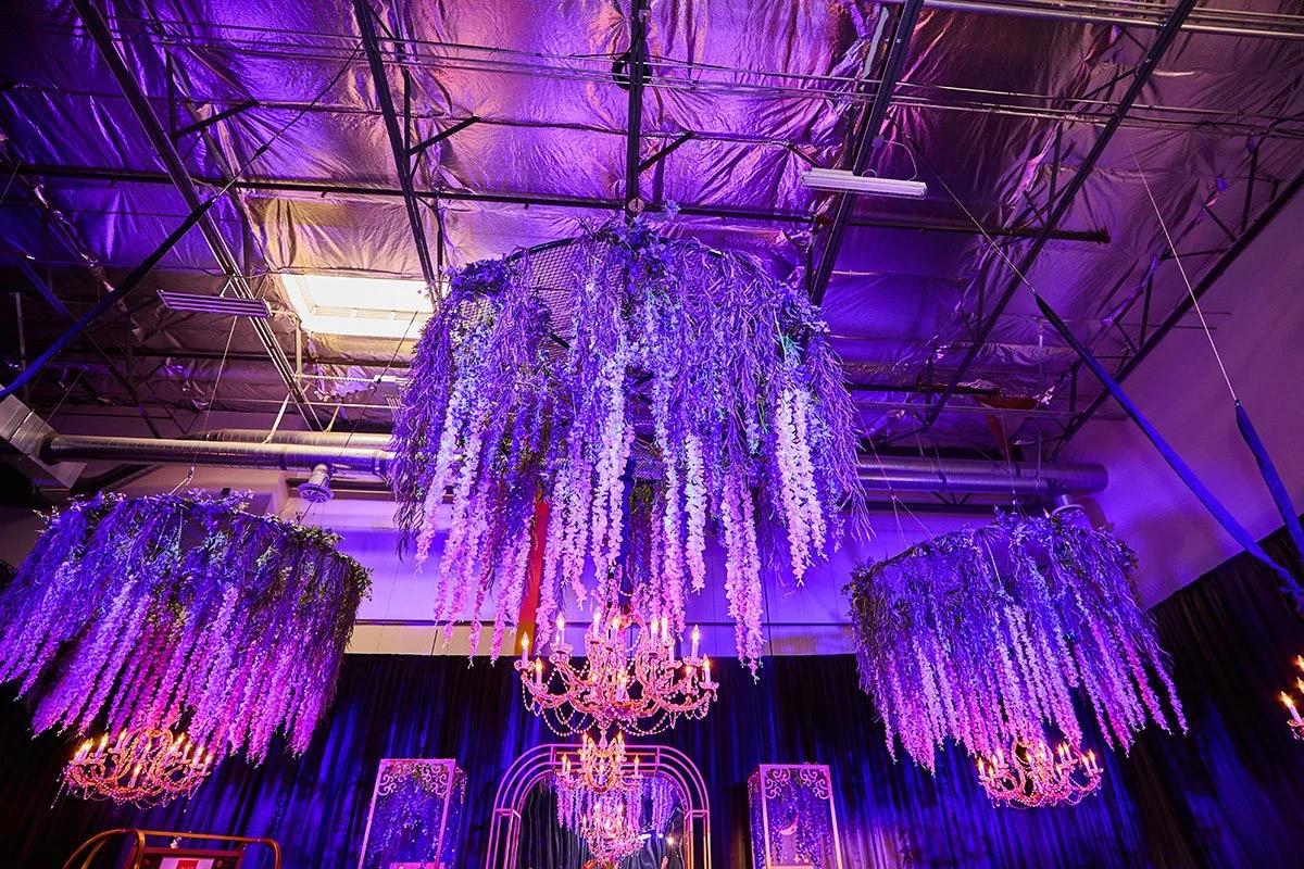 Elegant event venue decorated with large hanging floral arrangements in purple hues, crystal chandeliers, and dark drapery, illuminated with purple lighting.