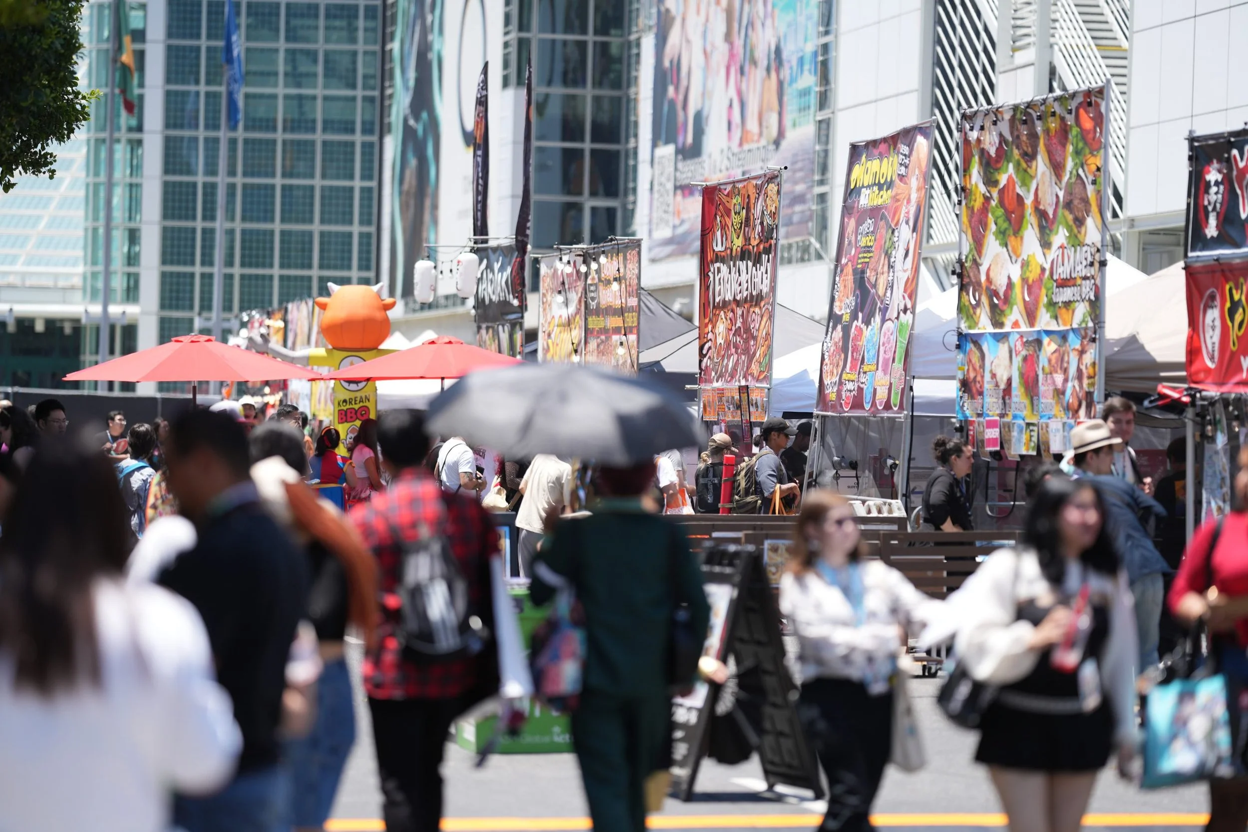 A crowded outdoor market with various booths selling food, with colorful banners, umbrellas, and a modern glass building in the background.