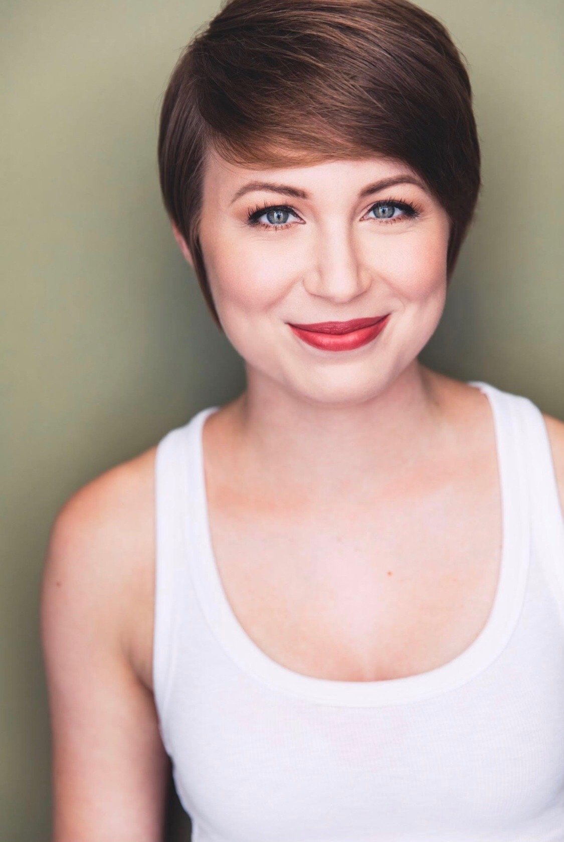 A woman with a short reddish-brown hairstyle, blue eyes, and red lipstick wearing a white tank top, smiling at the camera.