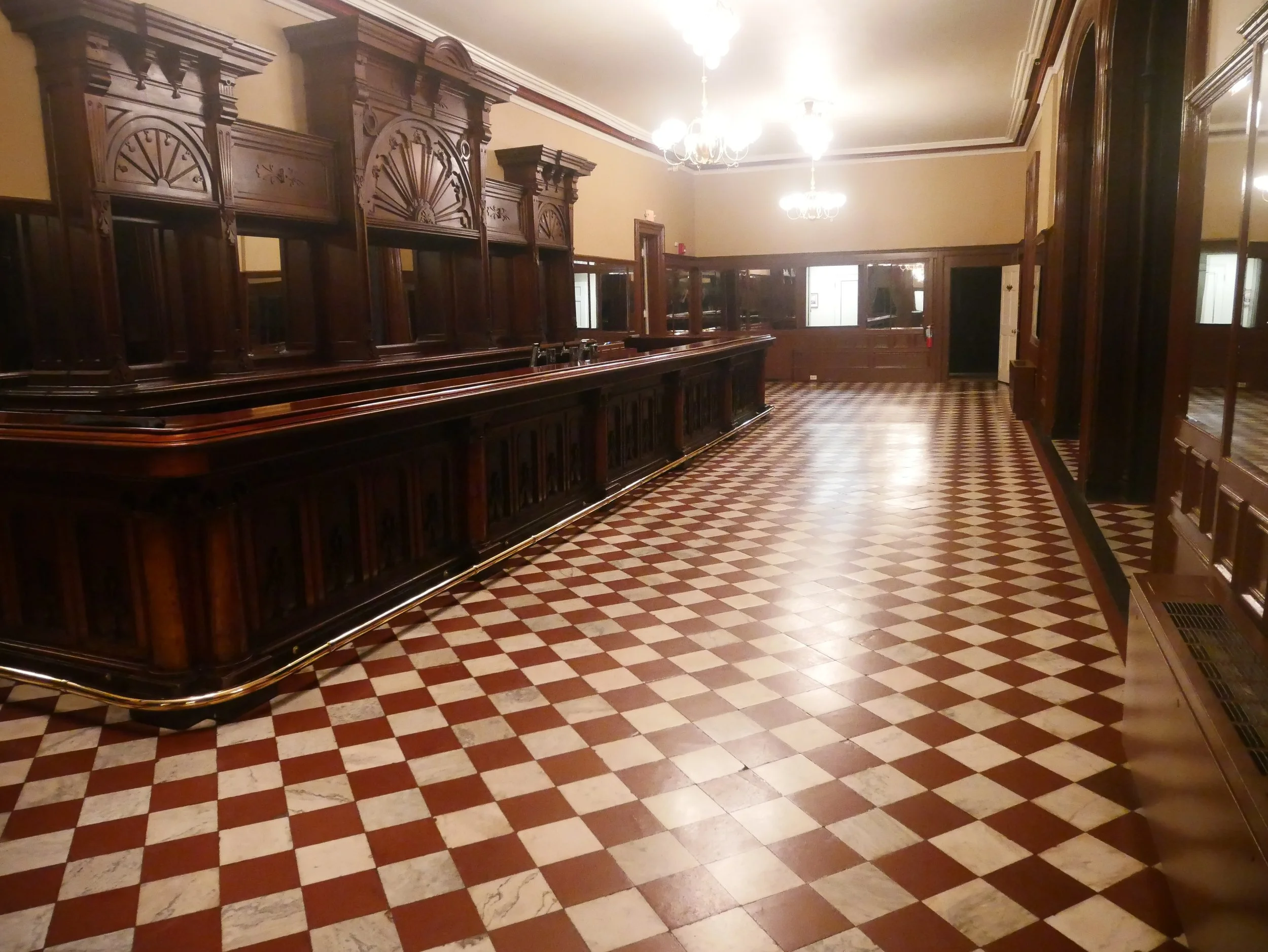 An empty, historic museum interior with dark wooden furnishings, chandeliers, and checkered tile flooring.