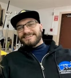 A smiling man with glasses, a beard, wearing a black baseball cap and a black hoodie with a blue patch, indoors with equipment and a door in the background.