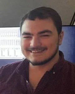 A young man with dark hair and a beard smiling at the camera, sitting indoors with a computer monitor in the background.