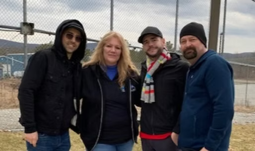 Four people standing outdoors near a chain-link fence with industrial structures in the background. It is cloudy, and they are dressed warmly.