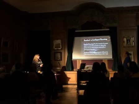 A woman presenting in front of a projected screen in a dimly lit room, with an audience seated and watching.