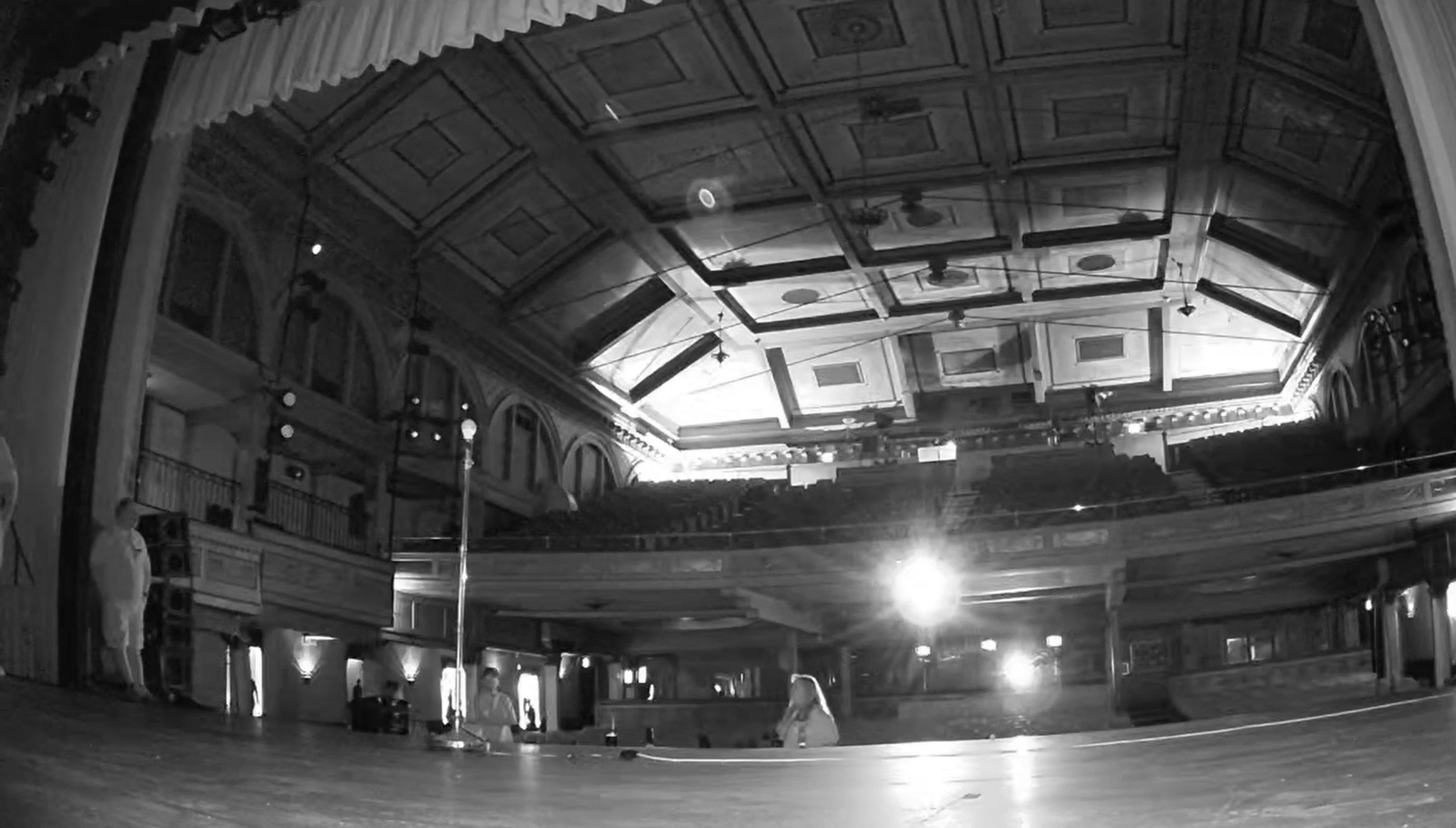 The image shows a large indoor theater or concert hall with a high, ornately decorated ceiling, and a stage area in the foreground. There are a few people standing around near the stage, and bright lights are illuminating parts of the hall.