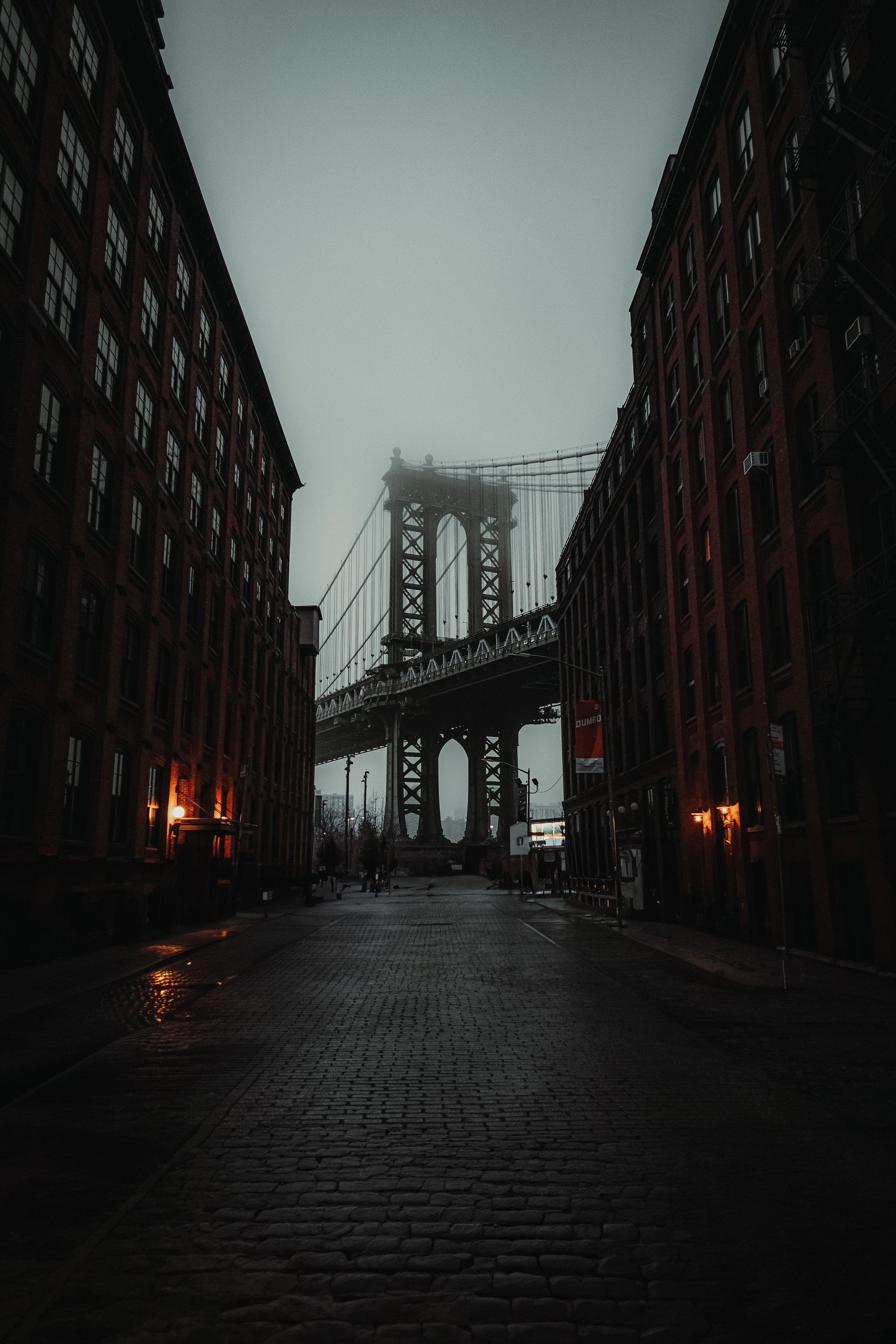 A foggy city street with cobblestone pavement, flanked by brick buildings, leading to the Brooklyn Bridge in the background.