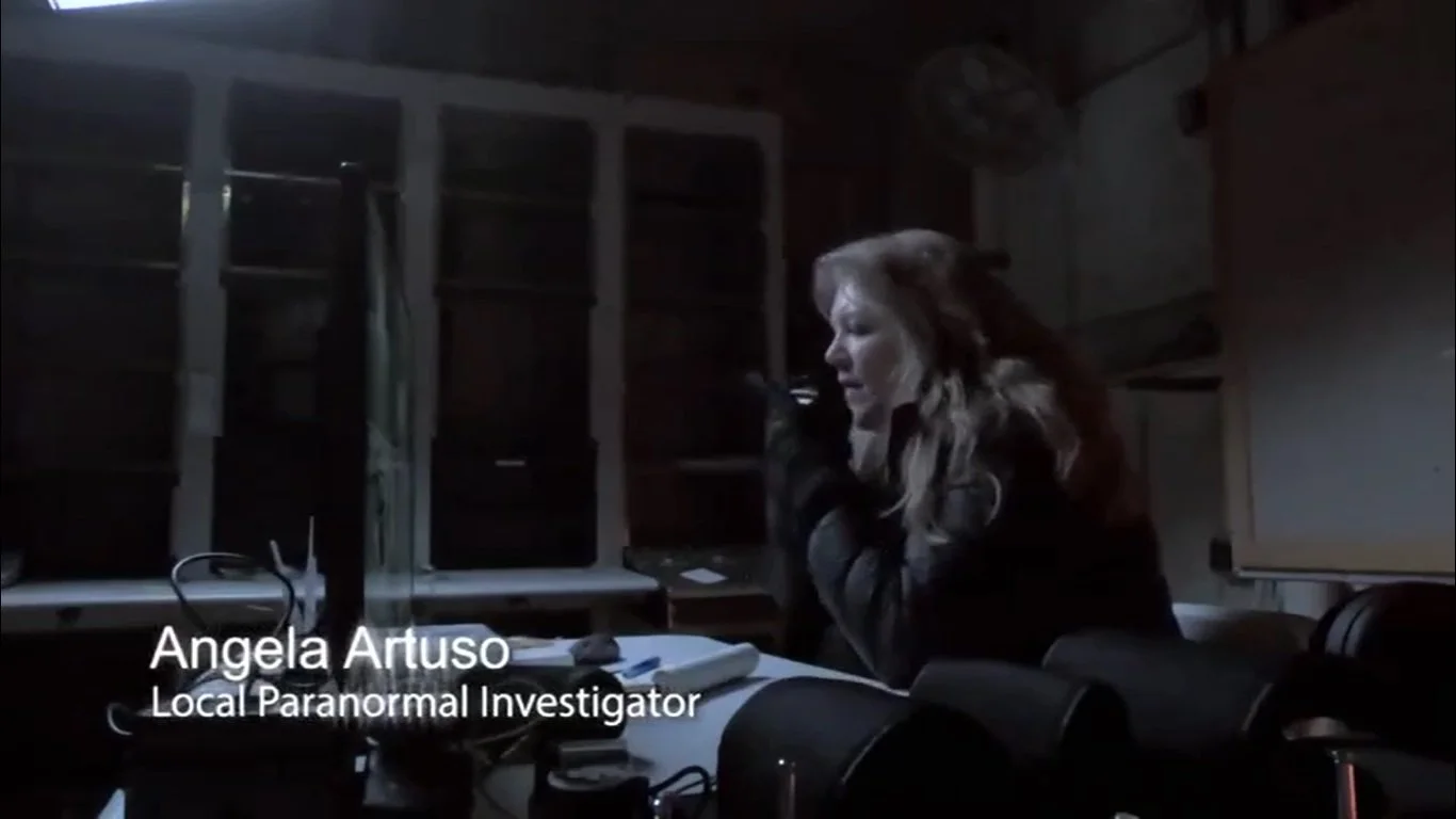 A woman with long wavy hair sitting at a table in a dark room, speaking into a camera. There is a computer monitor and some electronic equipment on the table. Text over the image reads, 'Angela Artuso, Local Paranormal Investigator.'