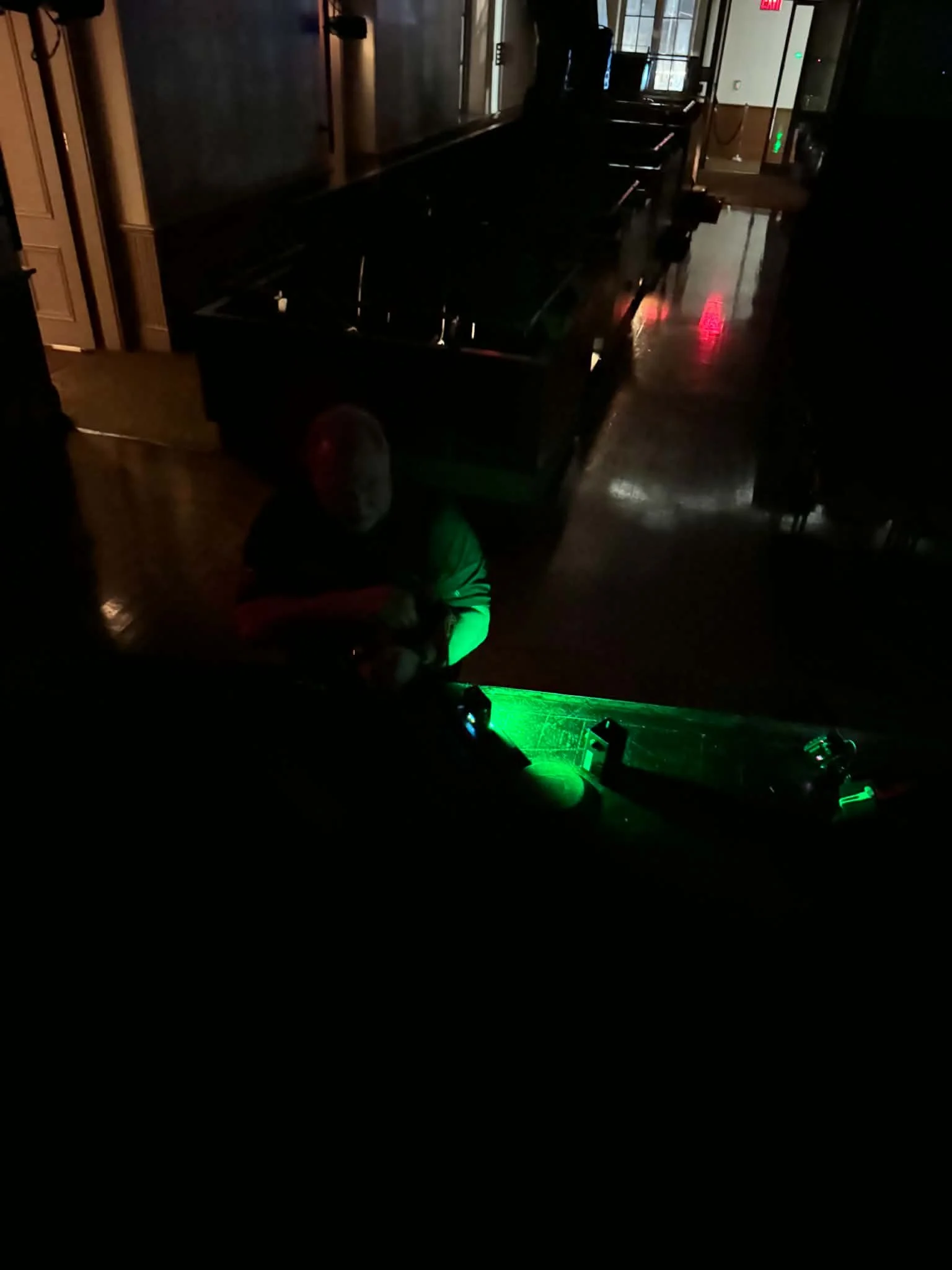 Dimly lit room with arcade game machine, person wearing green shirt and white cap playing, reflections of red and green lights on the wooden floor.