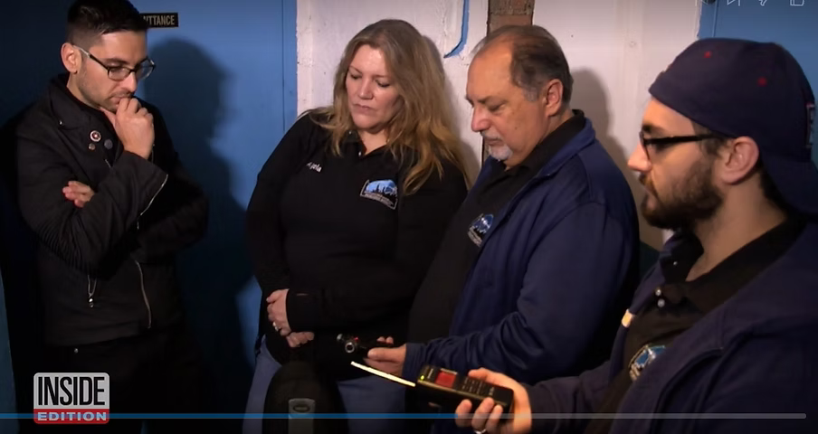 Four individuals are gathered together, looking at a handheld device, in an indoor setting with blue walls. They appear to be focused on the device, possibly analyzing or reviewing something.
