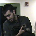 A young man with dark hair and beard looking down at a smartphone he is holding in his hands, standing in a room with green and white walls.