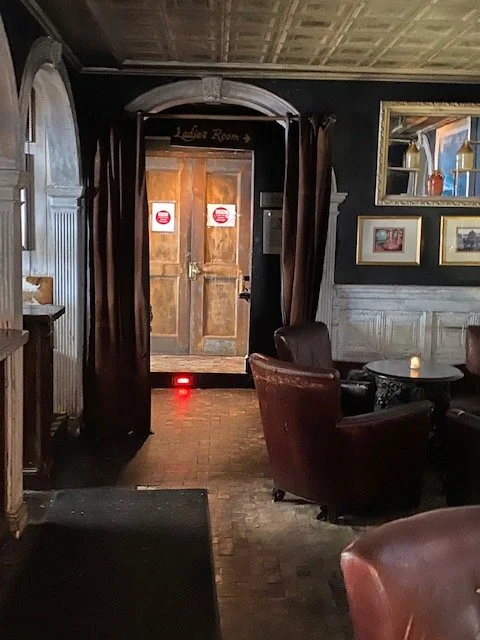Interior of a dimly lit historical restaurant or pub, with a wooden door in the background, framed pictures on the wall, and leather chairs around a small table.