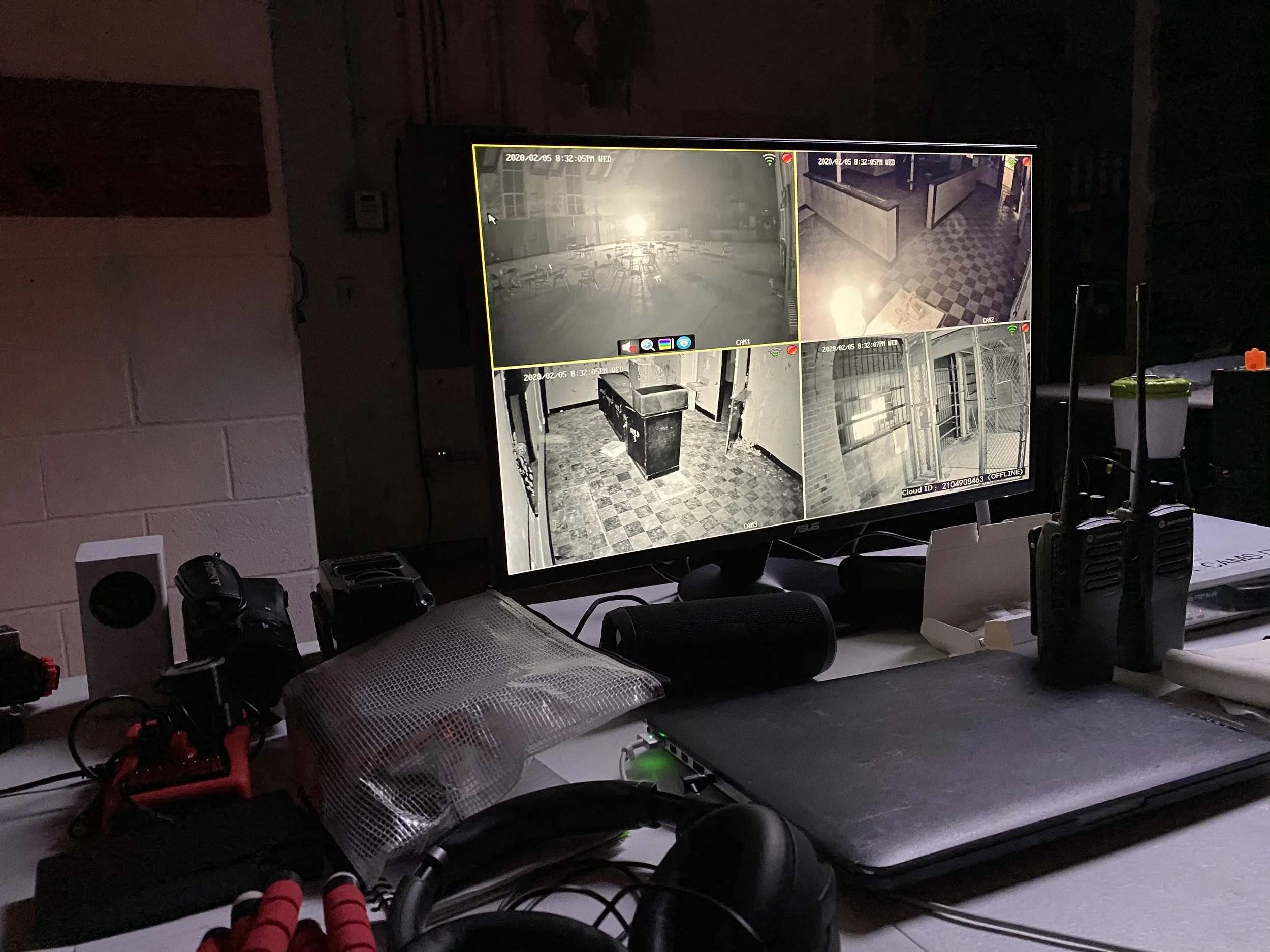 Security monitors showing surveillance footage of various indoor locations in a building, with equipment and cables on a desk below.