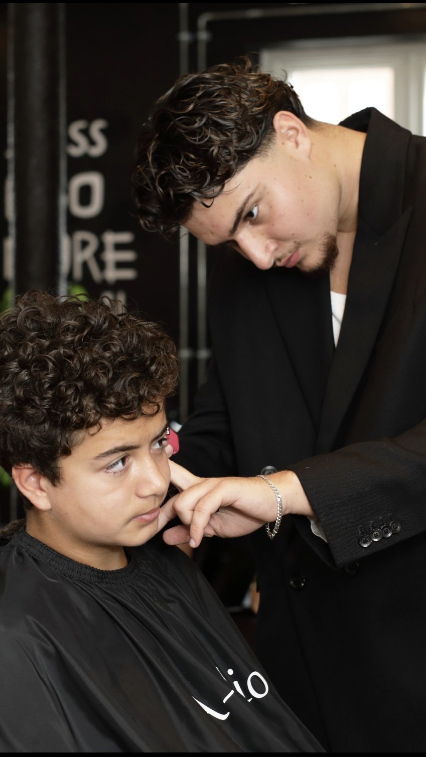 A young man with dark curly hair getting a haircut from a barber in a salon.