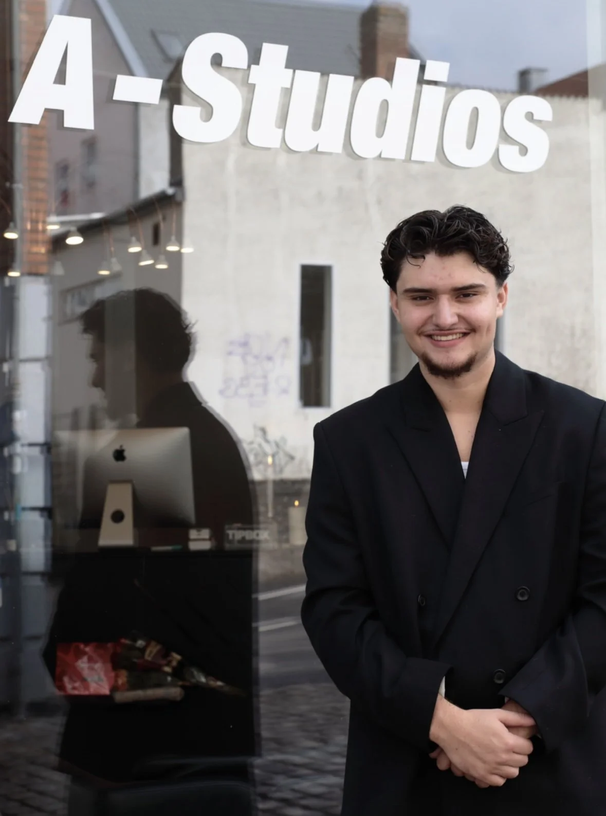 A young man with dark curly hair and a goatee smiling, dressed in a black blazer, standing outside a window with "A-Studios" written on it.