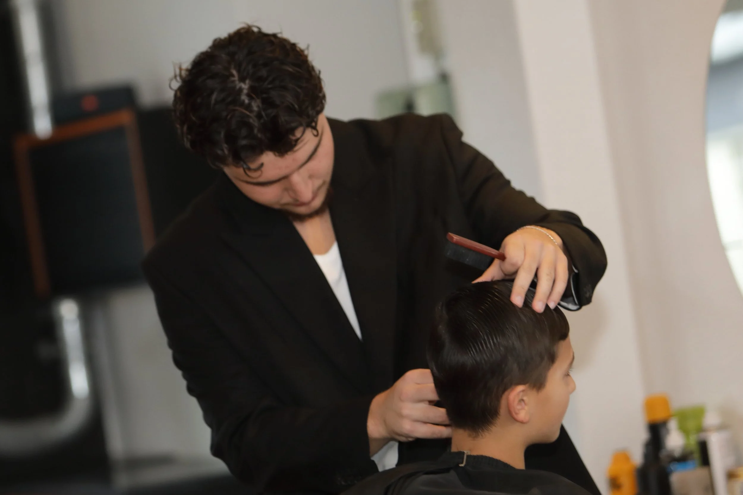 A hairstylist with curly hair and glasses cutting a client's hair at a salon.