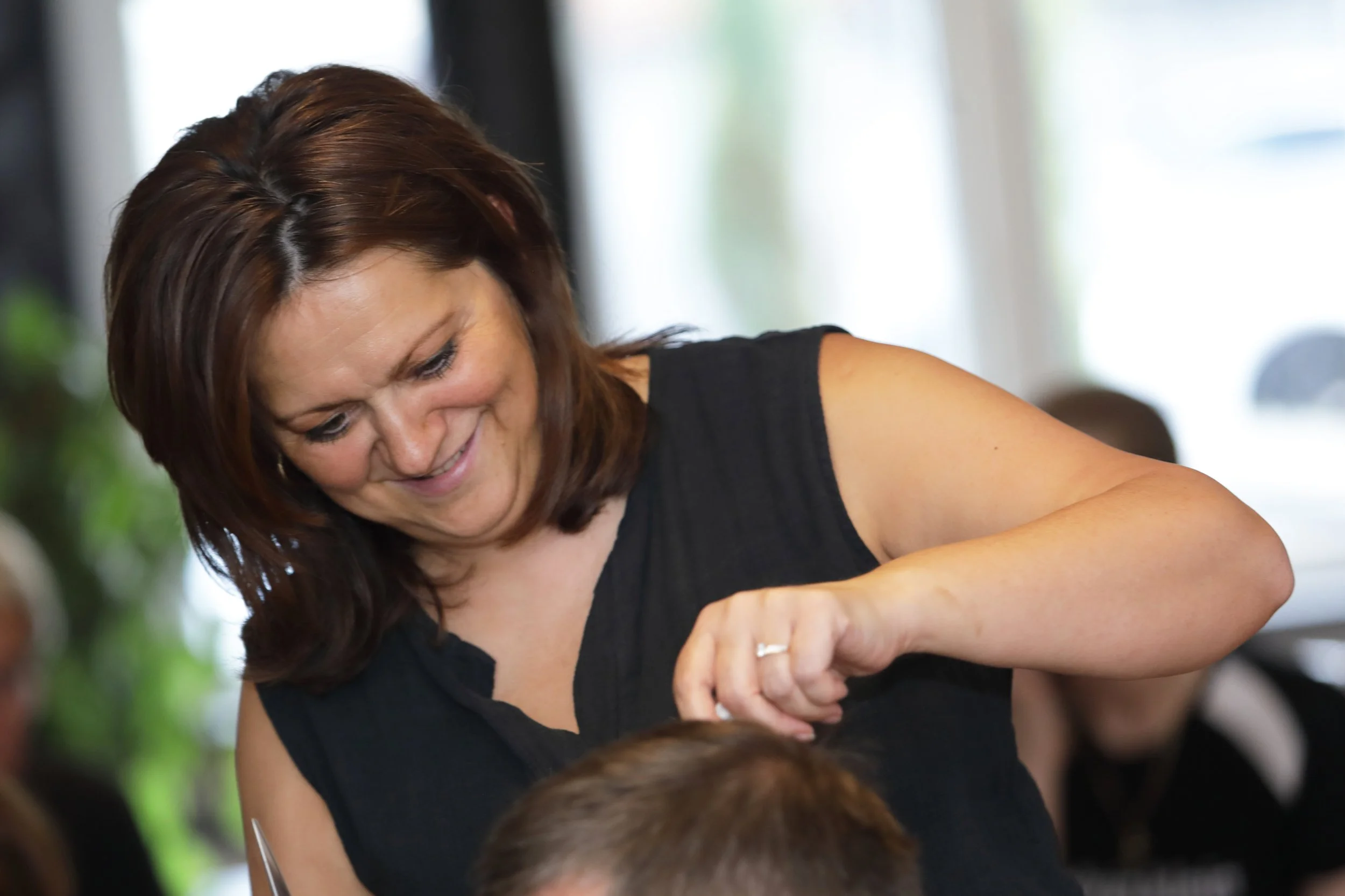 A woman with shoulder-length brown hair smiling and looking down, wearing a black top, engaging with children in an indoor setting.