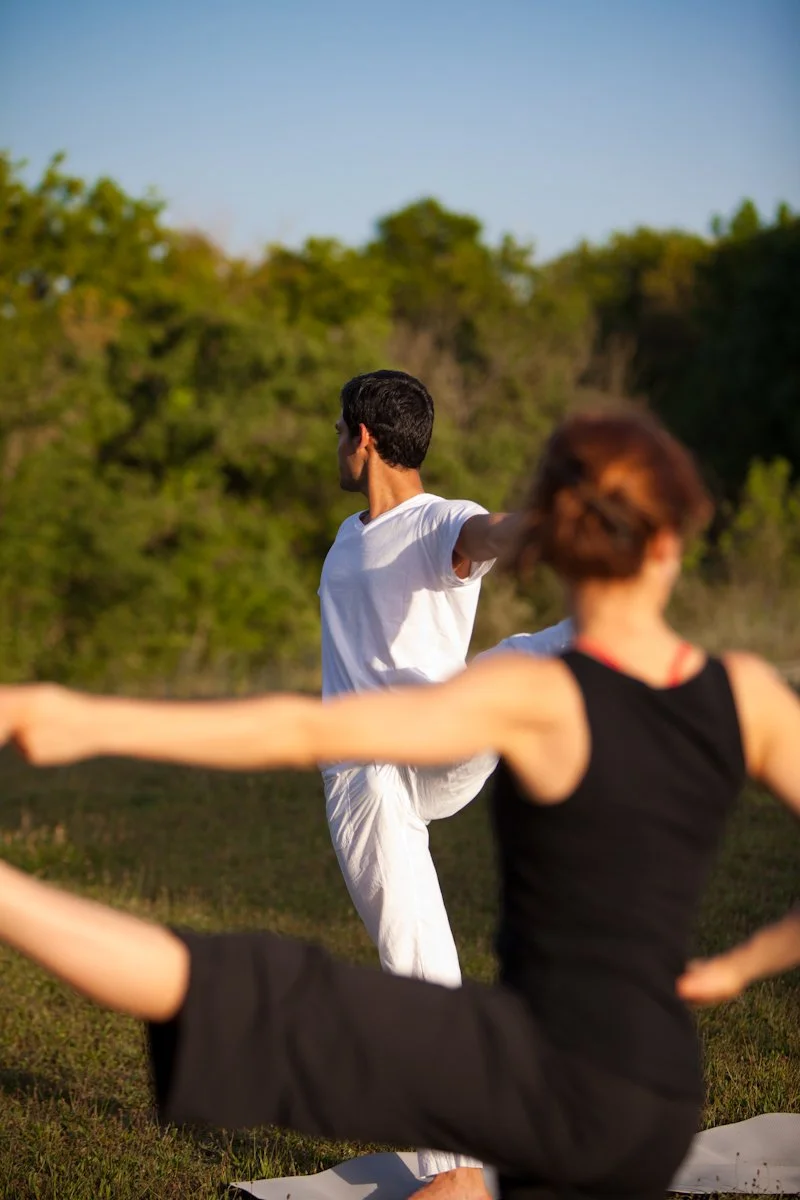 Personas realizando una práctica de yoga o meditación en un campo al aire libre, con árboles y cielo despejado en el fondo.
