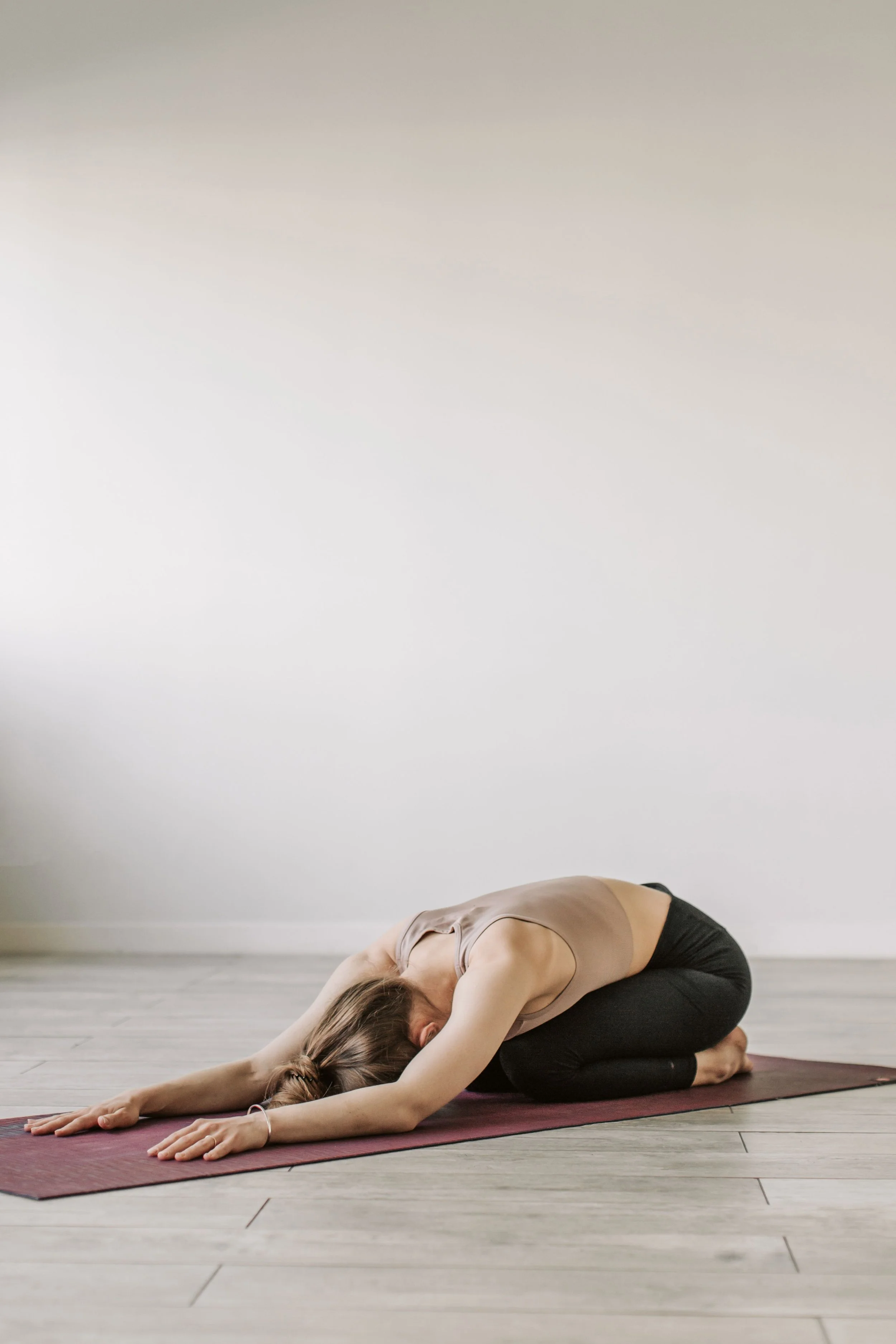Mujer haciendo yoga en posición de flexión hacia adelante en una esterilla en un espacio con paredes blancas y piso de madera.