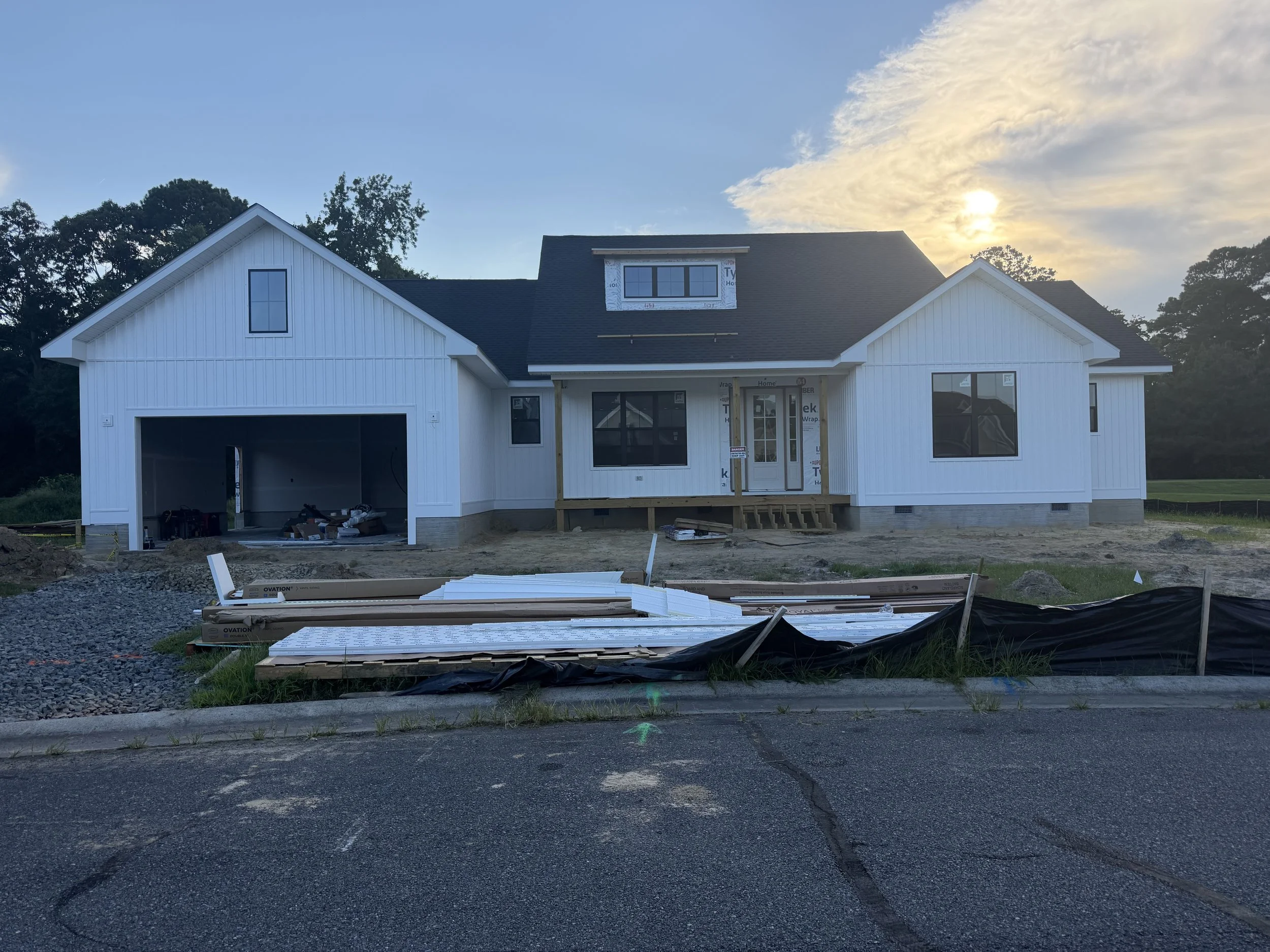A house under construction with white siding, a black roof, and a garage. The front porch is incomplete, with construction materials and tools, and the surrounding yard is unfinished with dirt and gravel. The sky is partly cloudy with the sun setting