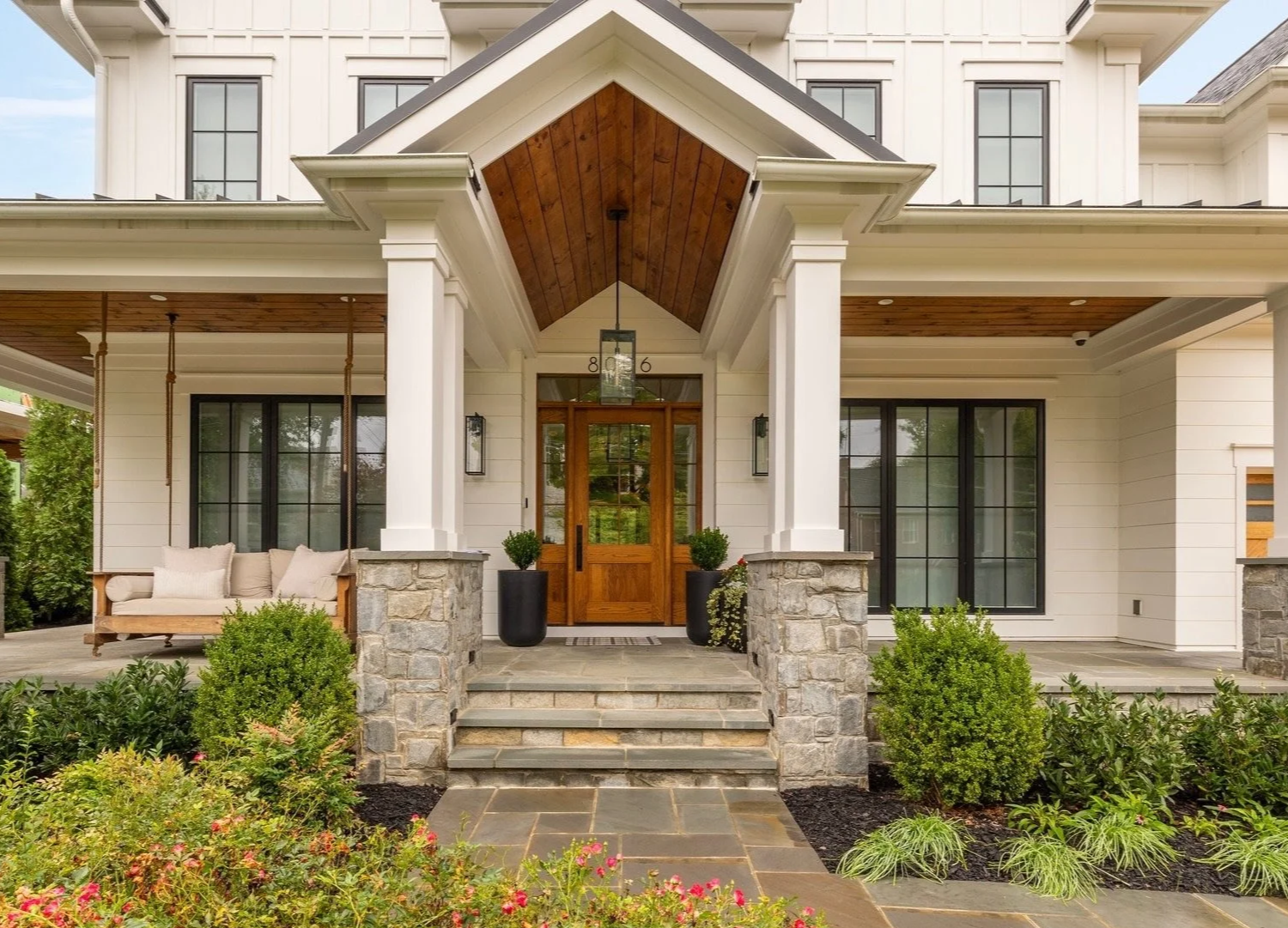 Front porch of a modern house with stone steps, white siding, large black window panes, a wooden front door, potted plants, and a bench with cushions, surrounded by landscaped bushes and flowers.