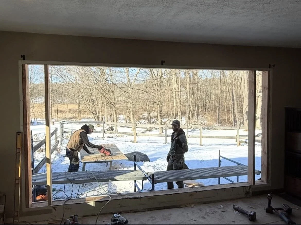 Two construction workers are working outside in snowy weather, installing or framing a large window in a building, with a wooden fence and trees in the background.