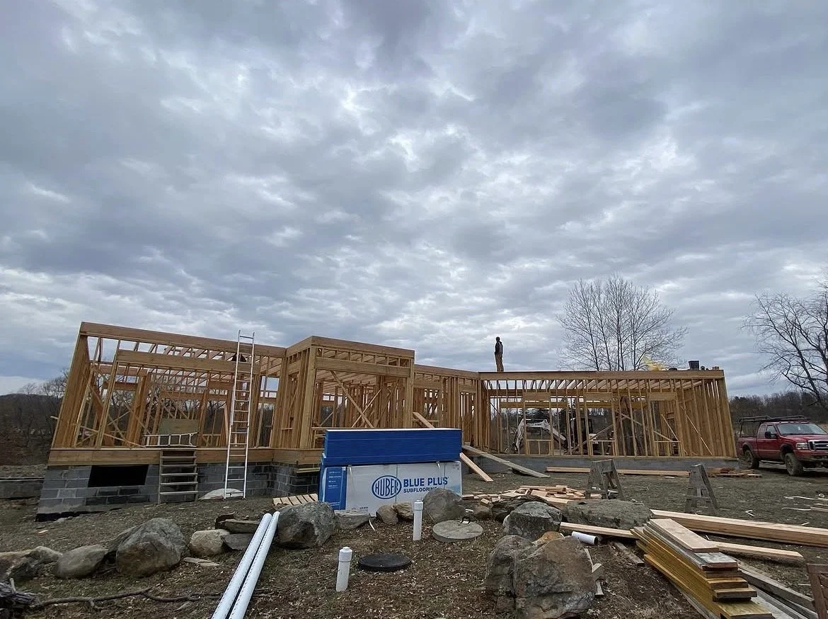 Construction site with a wooden framing house, a worker standing on top, and building materials scattered around under a cloudy sky.