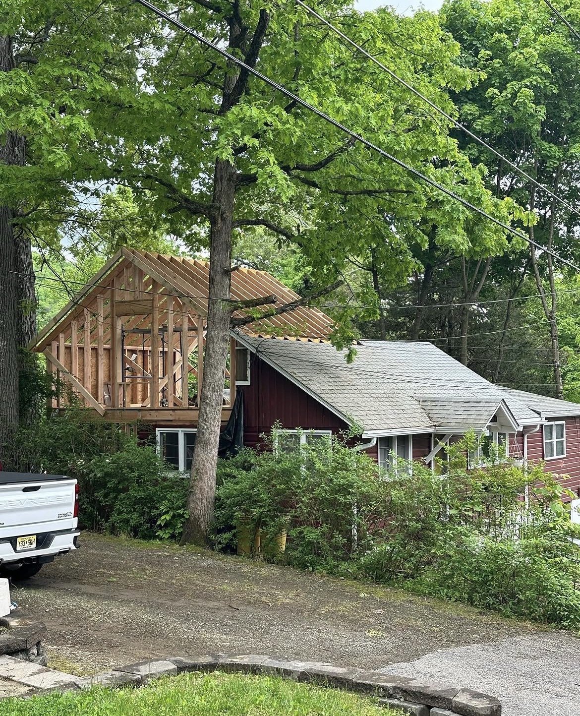 A house undergoing construction with a wooden framework for a second story, surrounded by green trees, with a white truck parked nearby.