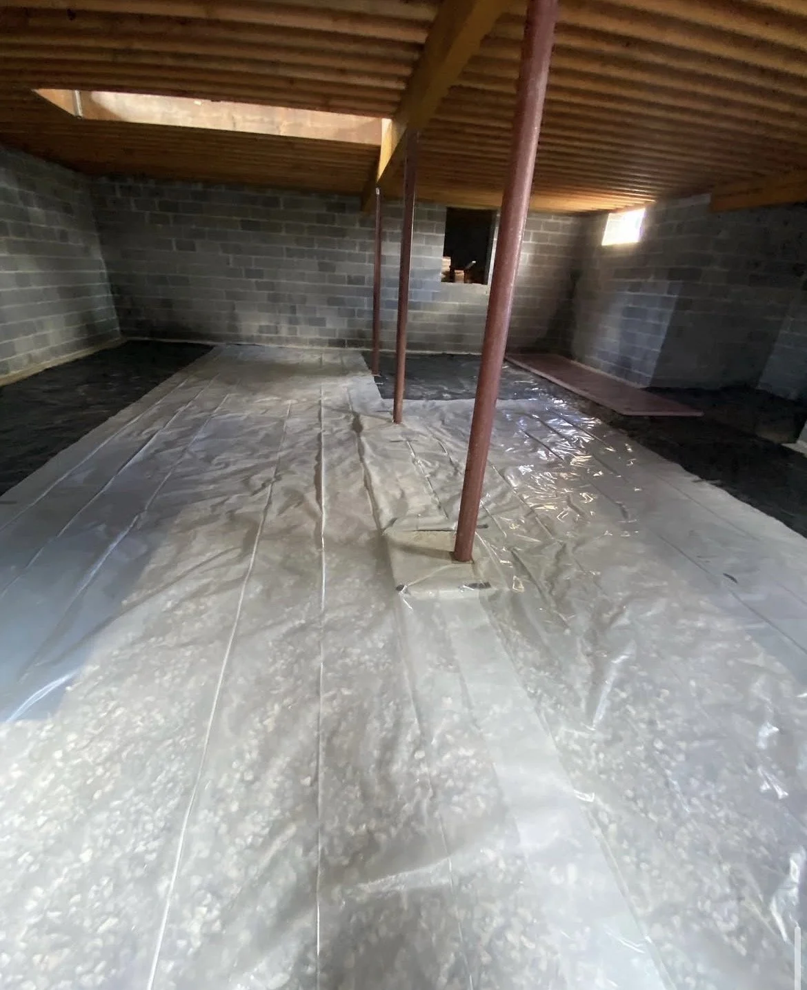Construction site with a floor covered in clear plastic sheeting, brick walls, and a wooden ceiling. Metal support poles are in the center of the room, and there is a small window on the wall.