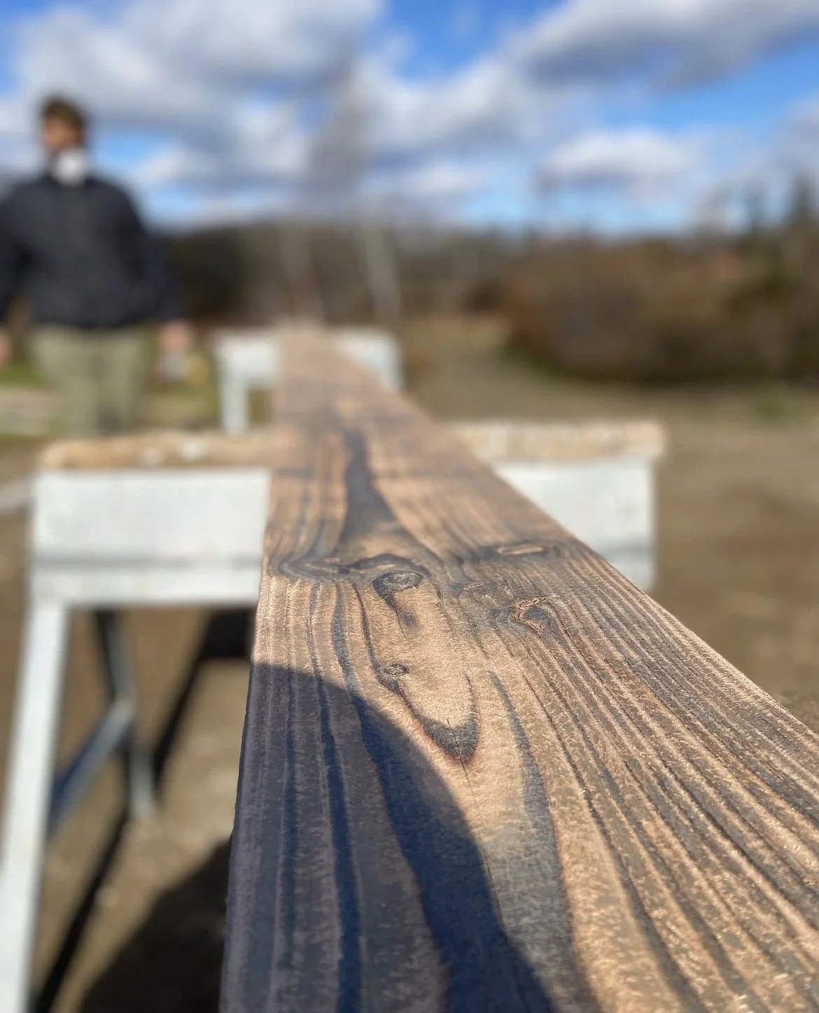 Close-up of a wooden plank on a table with a person in the background outdoors under a cloudy sky.