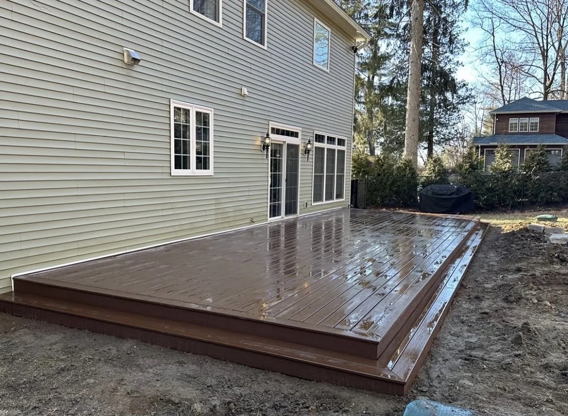 Newly built wooden deck attached to the back of a house with beige siding, multiple windows, and a sliding glass door. The deck is wet and shiny from rain, with an overgrown yard and other houses visible in the background.
