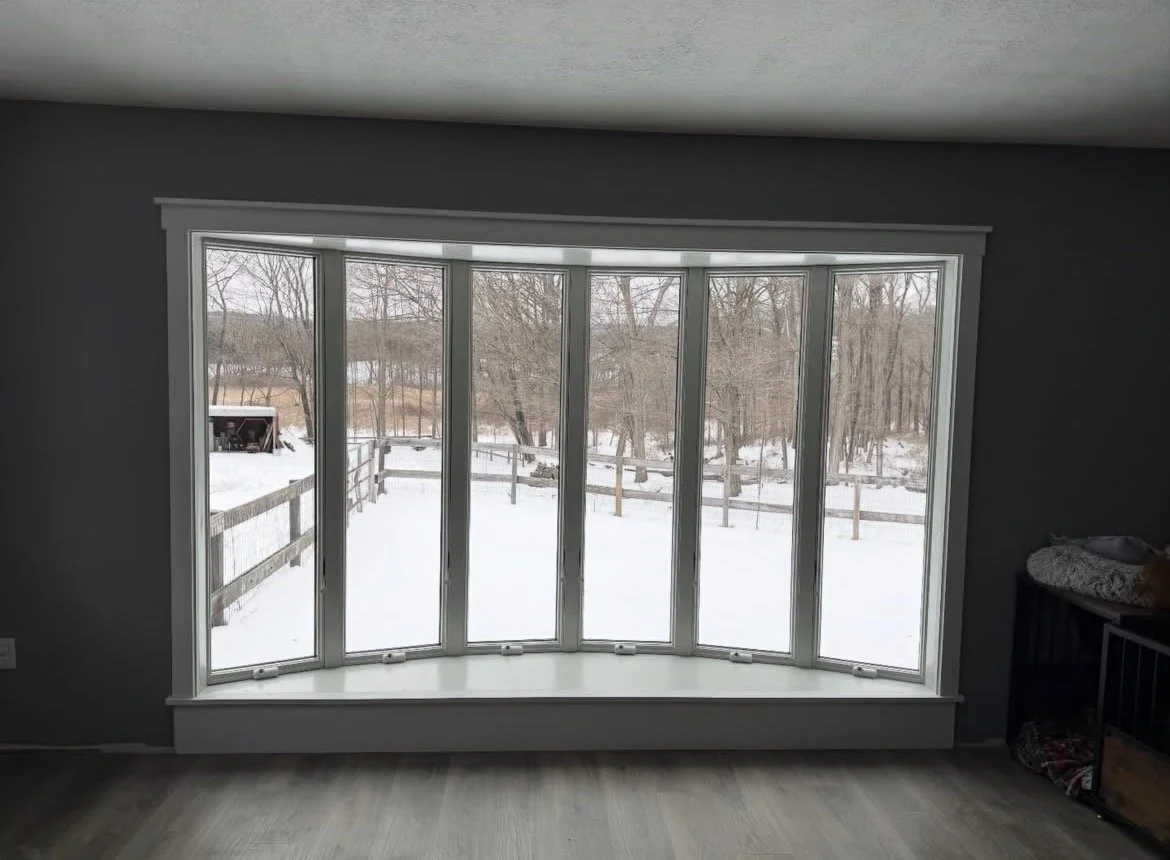 View of a snowy backyard seen through a large bay window in a room with dark walls and light-colored wood flooring.