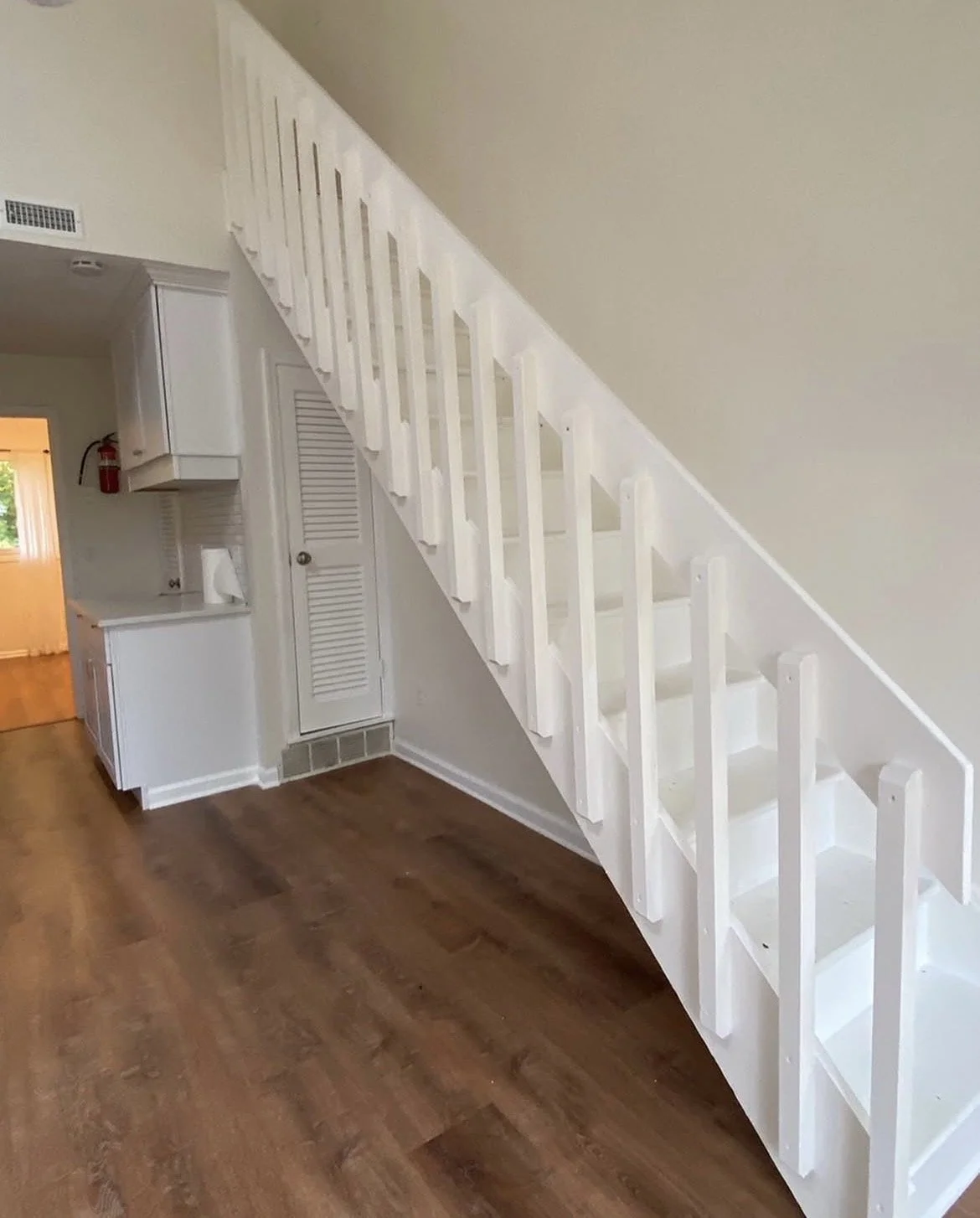 Interior view of a room with a white staircase leading upstairs, wooden flooring, and kitchen area in the background.