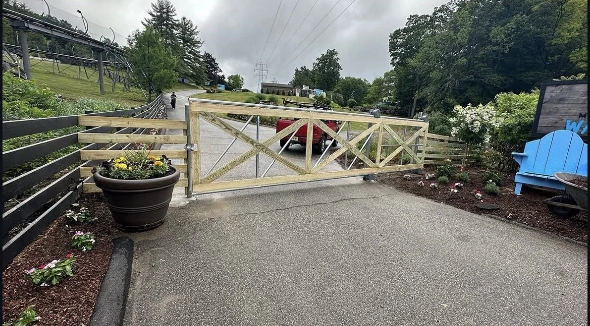 Wooden gate blocking a driveway with a red truck parked behind it, surrounded by flower beds and outdoor seating, under an overcast sky.