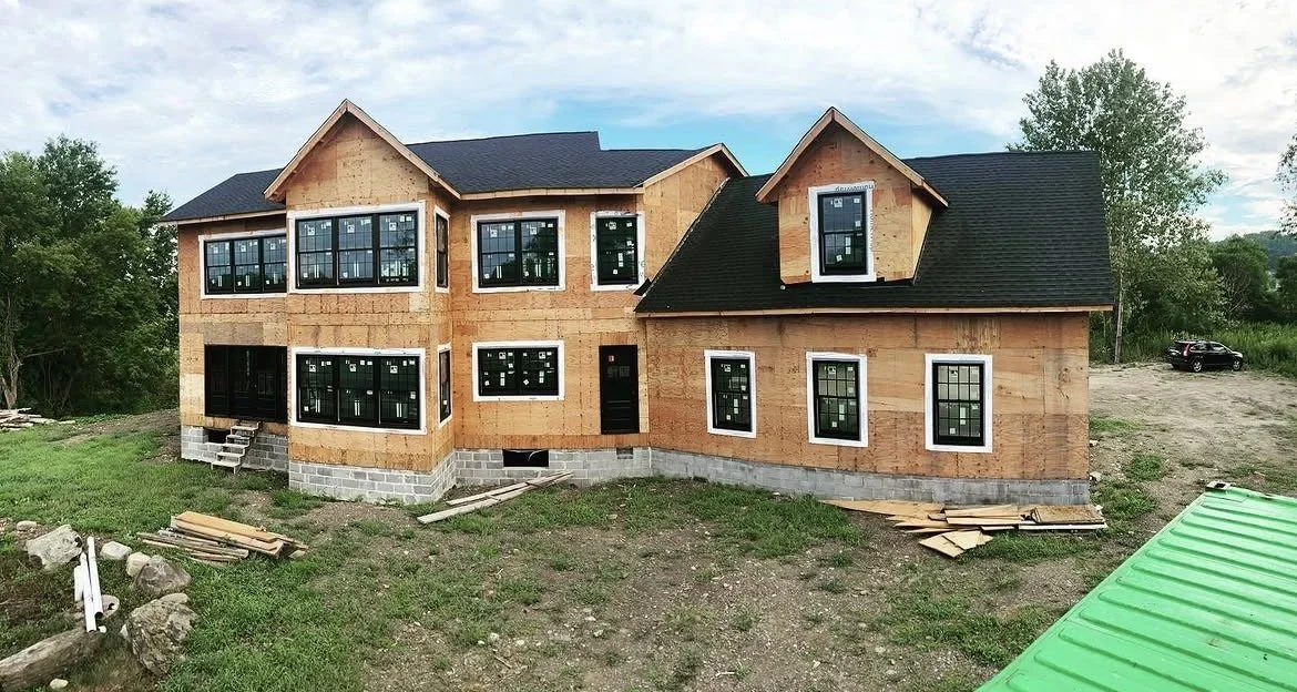 A house under construction with wooden framing, multiple large windows, and a dark shingled roof, surrounded by grassy land and trees.