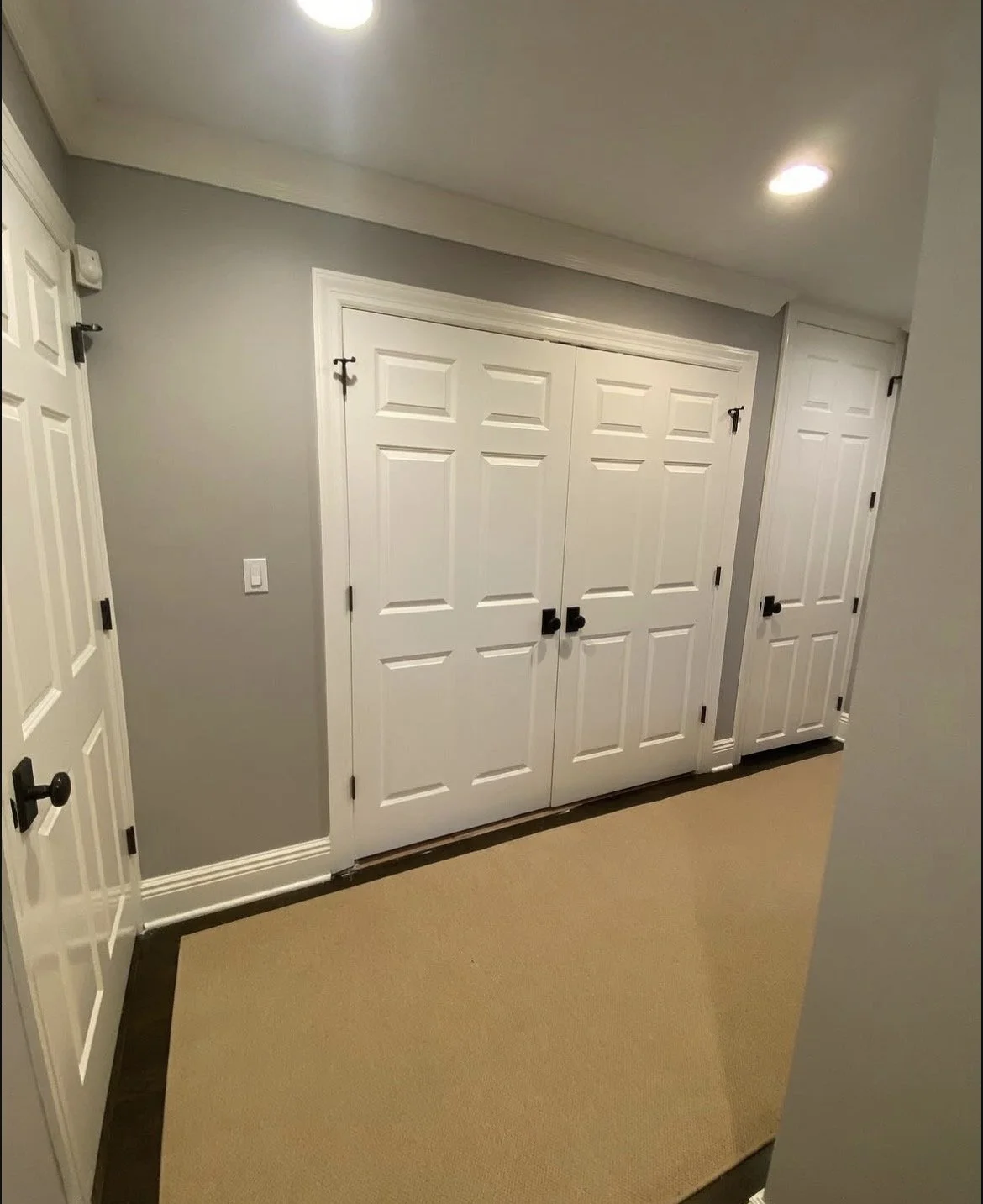 A hallway with gray walls, white trim, and four white doors with black knobs, two of which are sliding pocket doors, and a beige and dark brown rug on the floor.