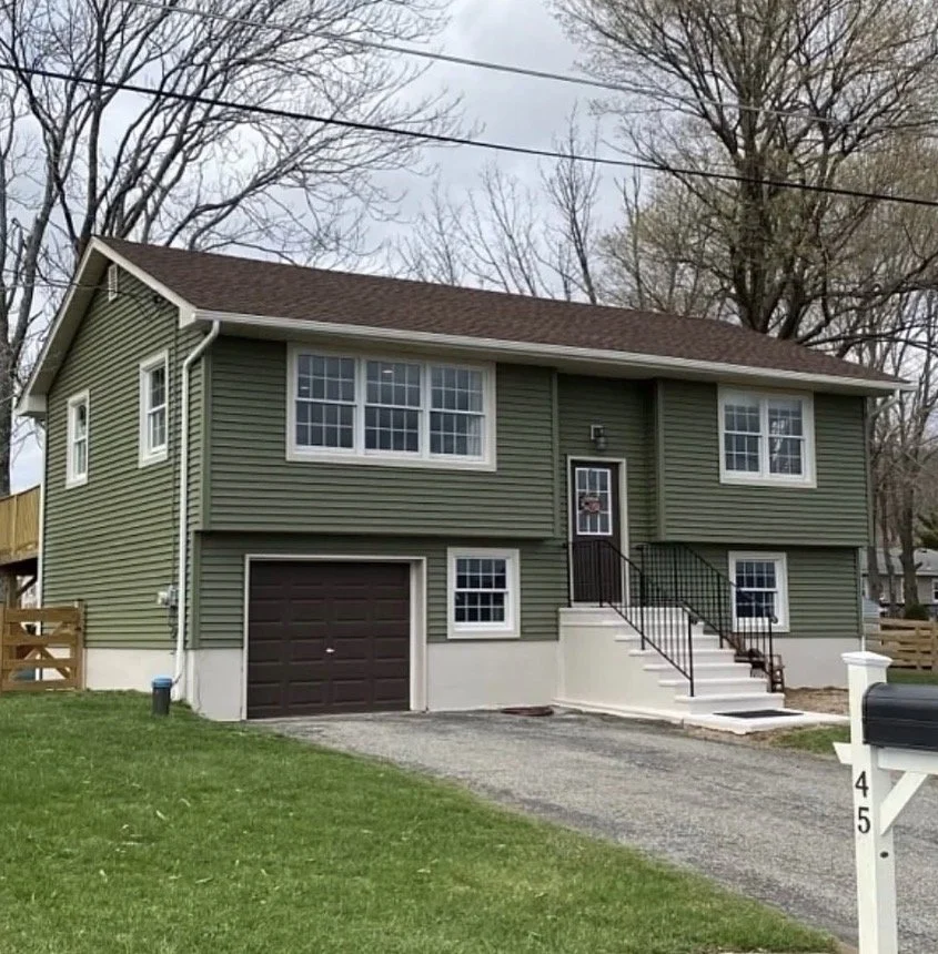 A two-story house with green siding, white trim, and a brown front door. The house has a one-car garage with a brown door. There are stairs leading up to the front door and multiple windows on both floors. The front yard has green grass and a gravel driveway, with a mailbox numbered 45 in the foreground.