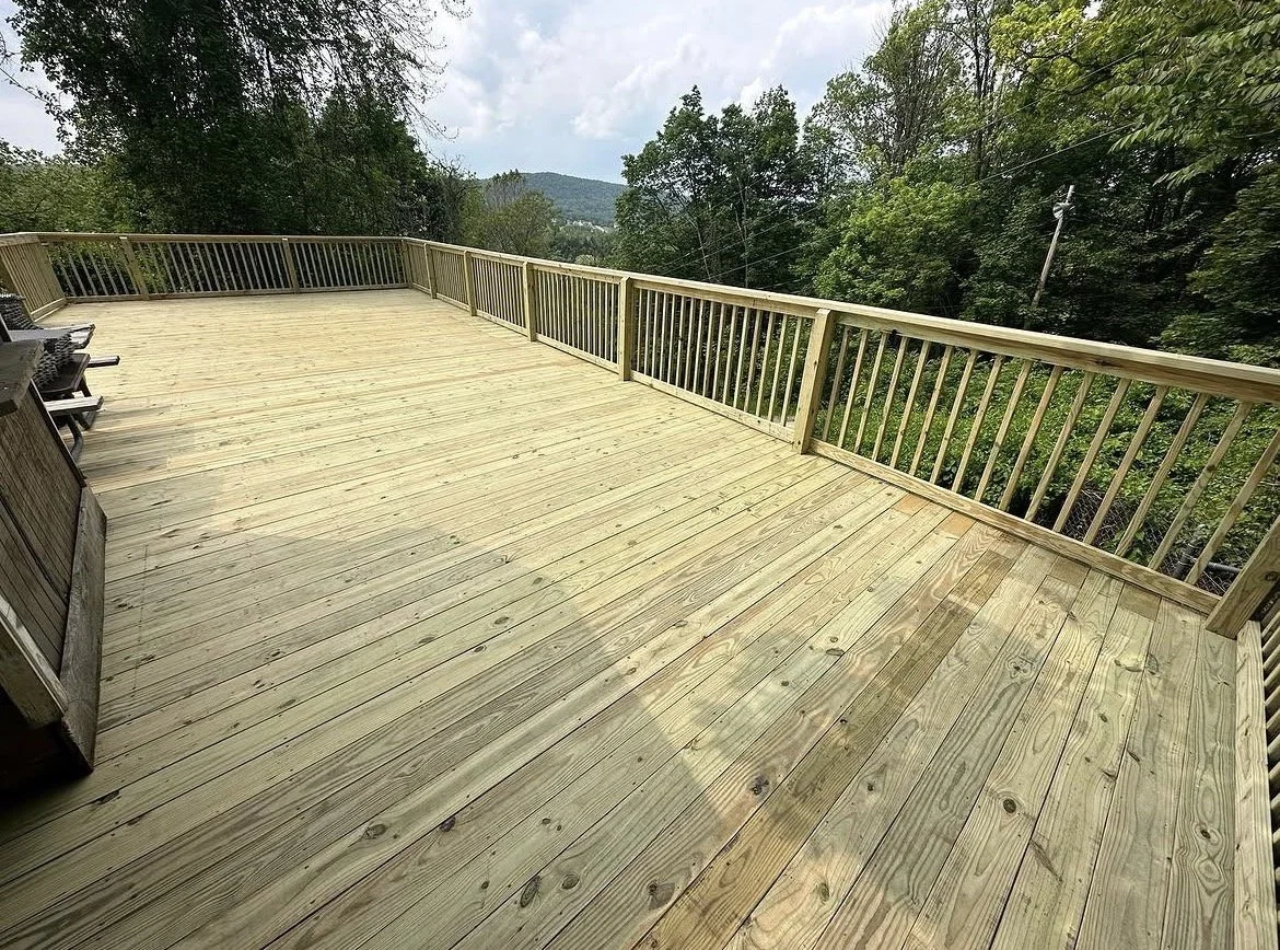 A spacious wooden deck with a railing, surrounded by trees and a distant view of hills under a cloudy sky.