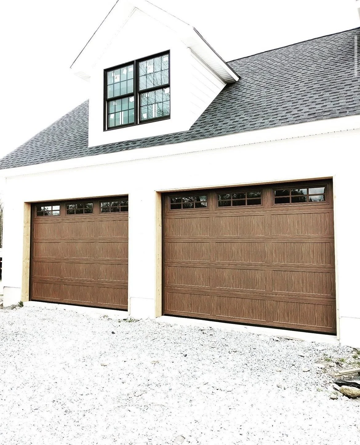 Photo of a modern house with two brown garage doors and a small dormer window with black framing on white siding in a suburban neighborhood.