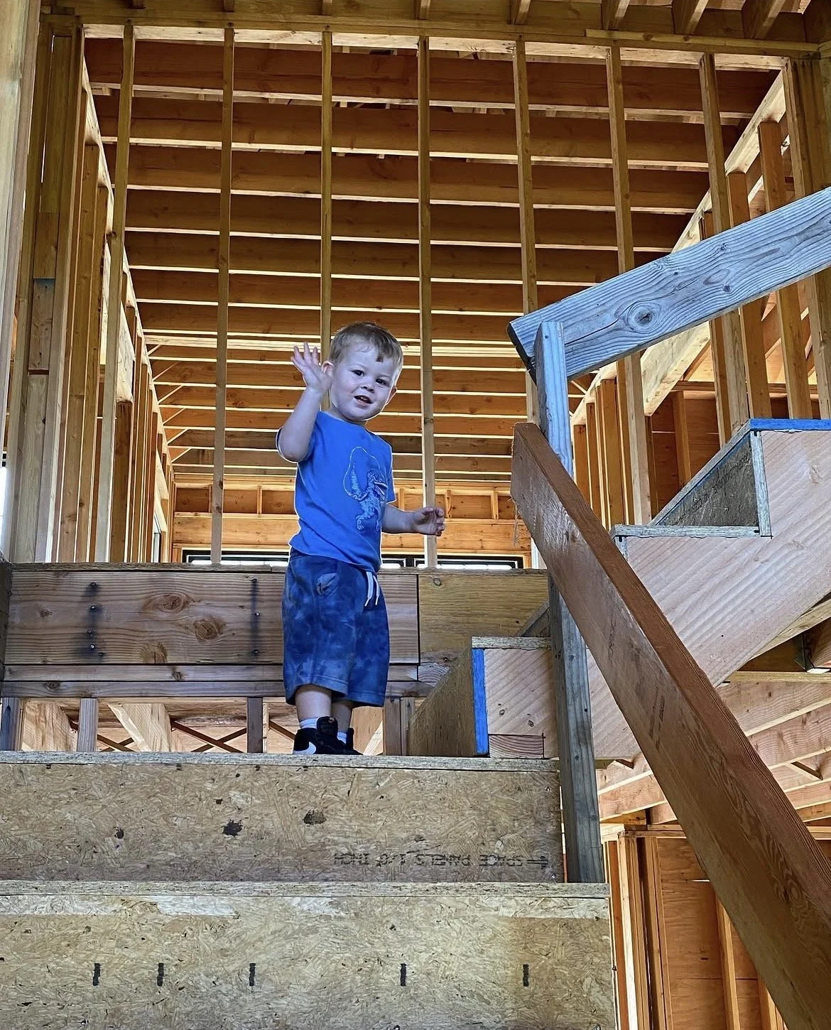 A young boy waves while standing on a partially constructed wooden staircase inside a house under construction.